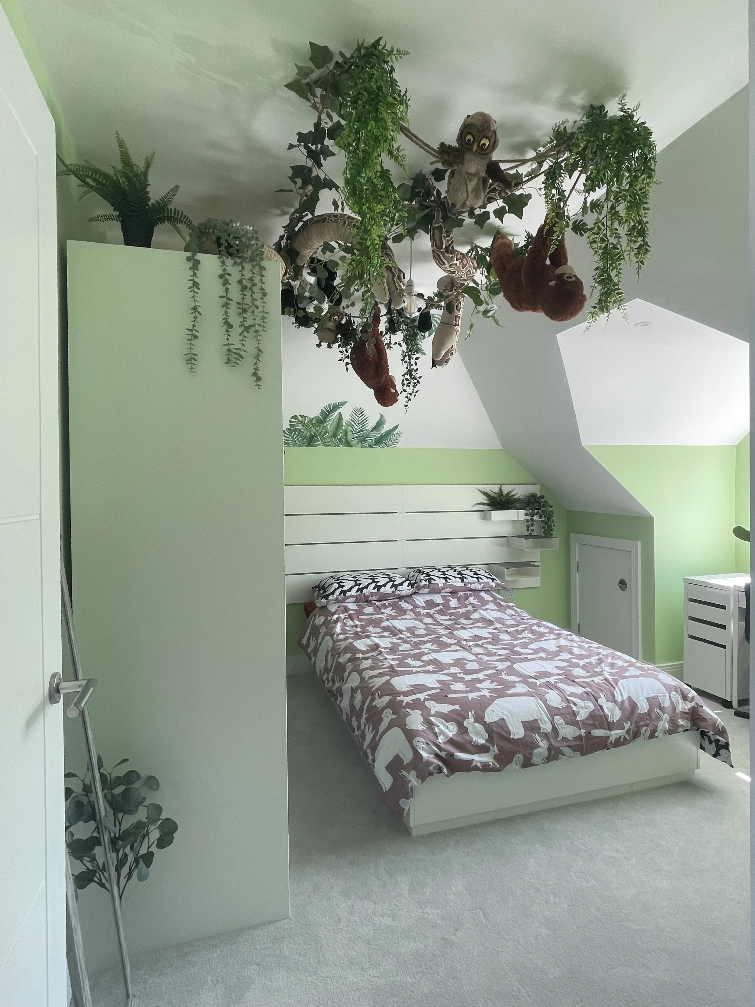 A bedroom with light green walls, a white bed with animal-themed bedding, and a ceiling decorated with plush animals and hanging greenery.