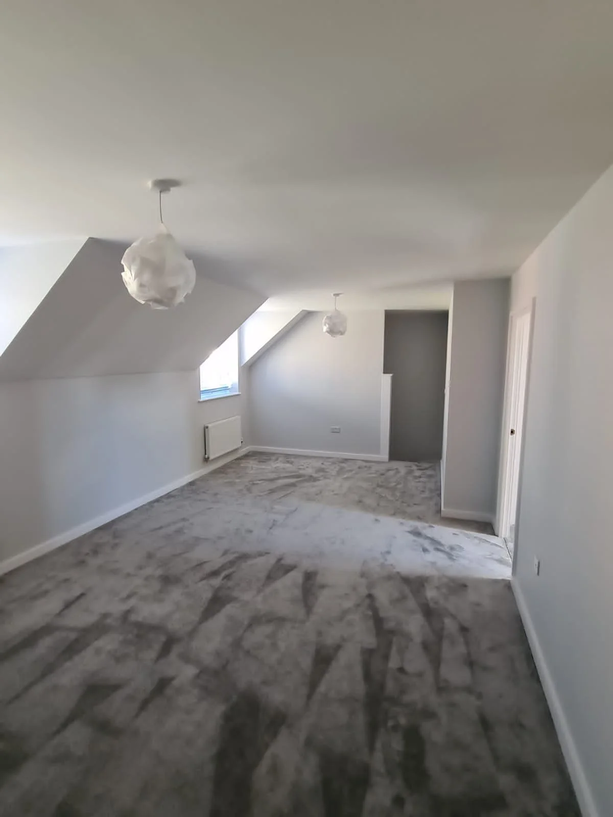 Empty, freshly renovated attic room with white walls, sloped ceilings, and a gray carpet.