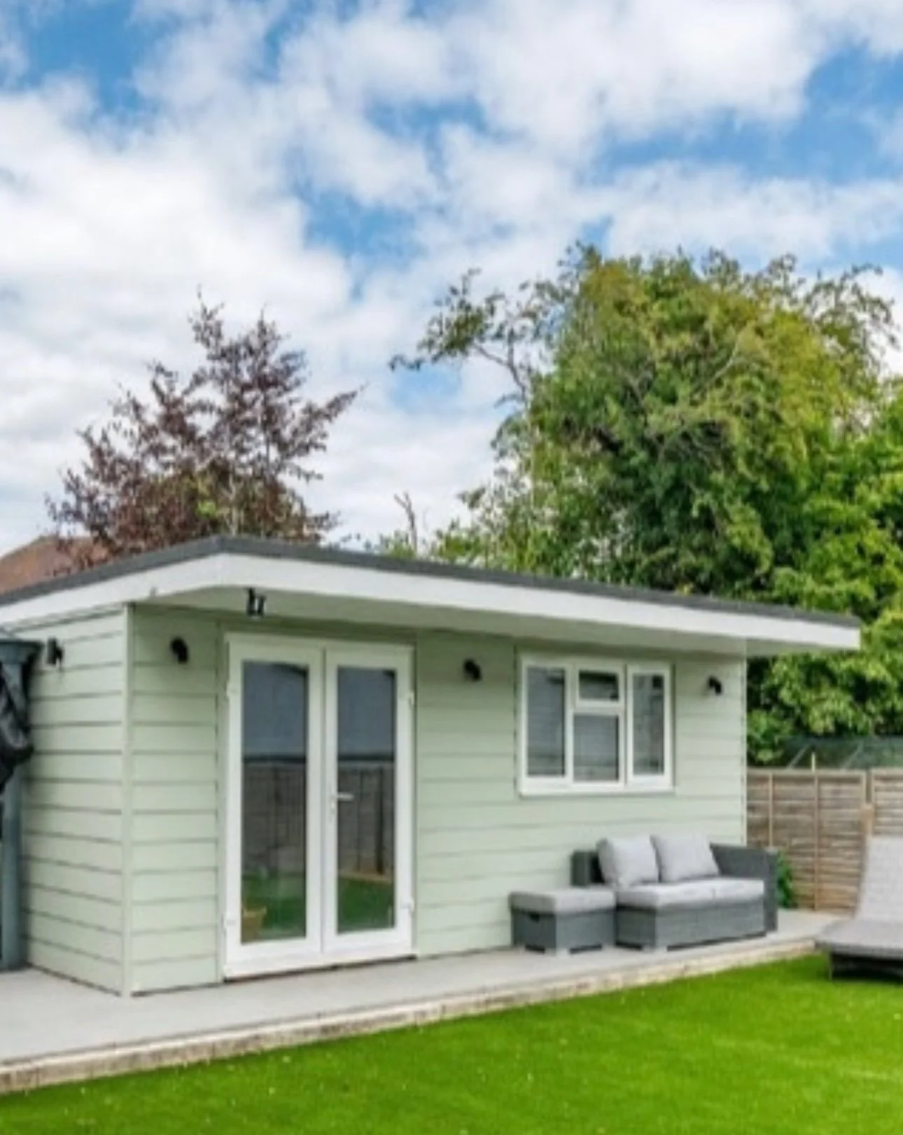 Backyard with a light green shed, outdoor seating, green grass, and trees under a partly cloudy sky.