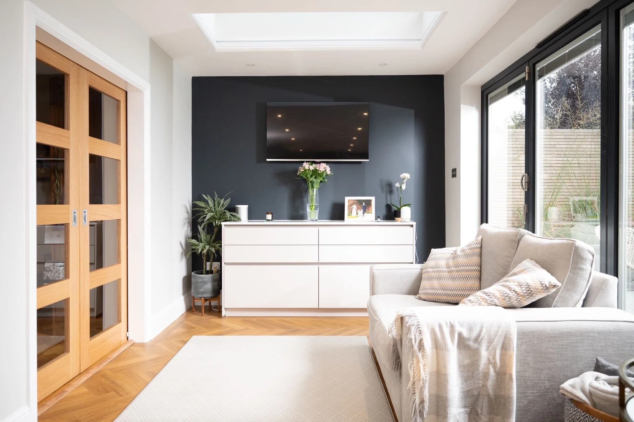 Modern living room with beige sofa, black accent wall, white sideboard with plants and photo frame, glass sliding door, and a wall-mounted flat-screen TV.