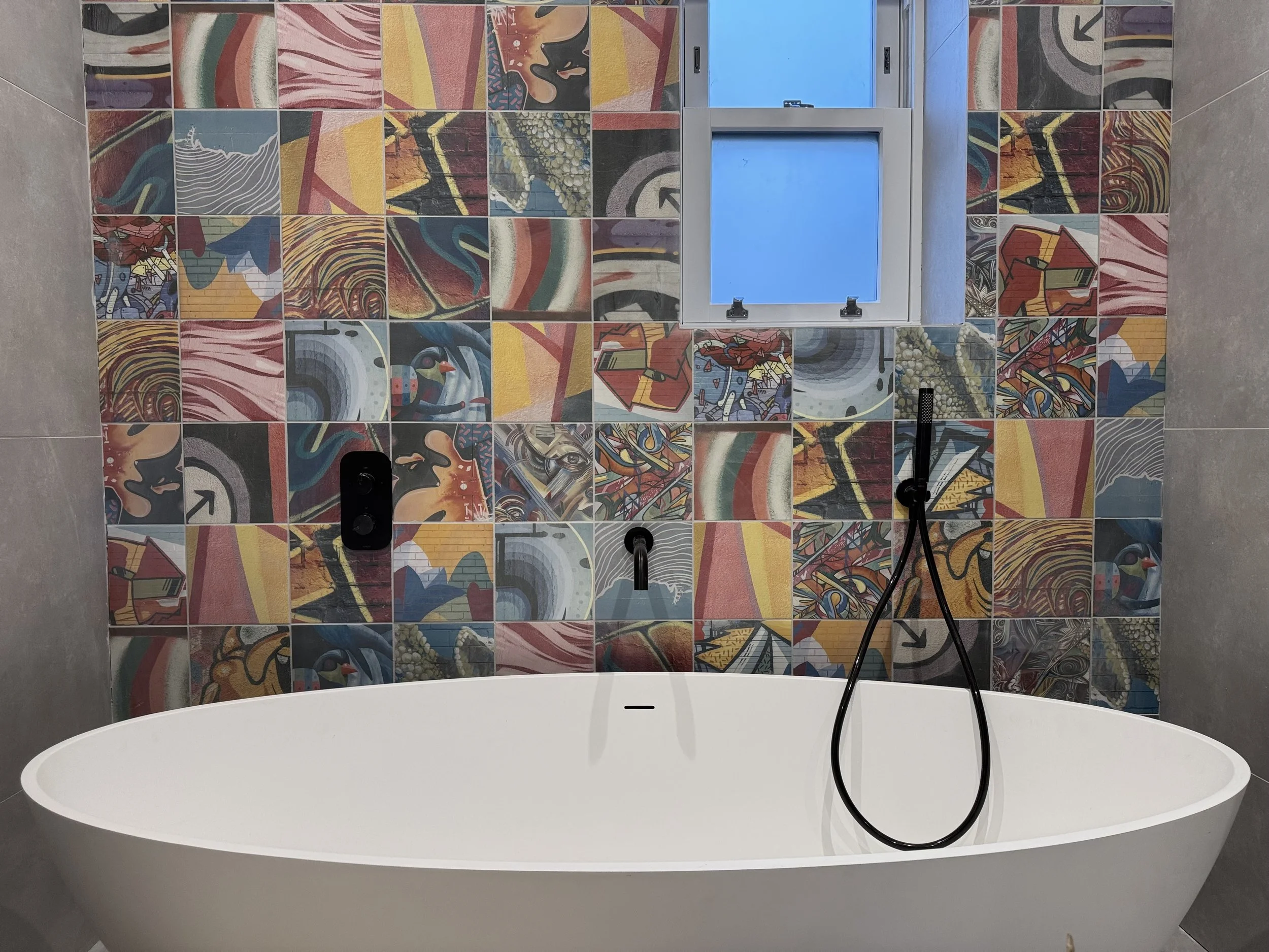 Modern bathroom with a freestanding white bathtub against a wall covered in colorful abstract patterned tiles. There is a window above the tub, a black shower head hanging from a hose, and black fixtures on the wall.