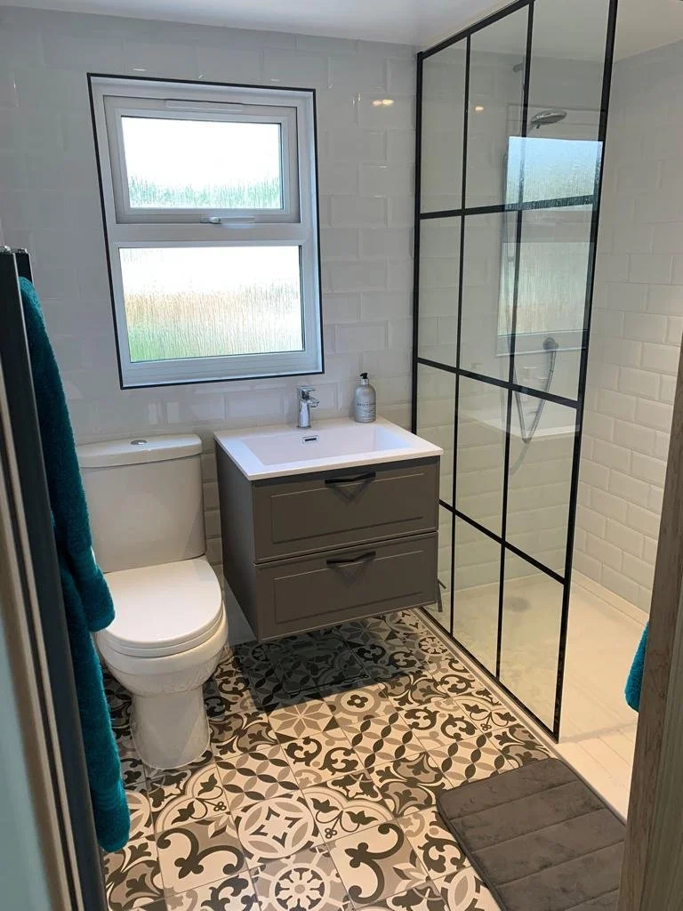 Modern bathroom with patterned floor tiles, a white toilet, a gray vanity with a white sink, a black-framed glass shower enclosure, and a window with frosted glass.