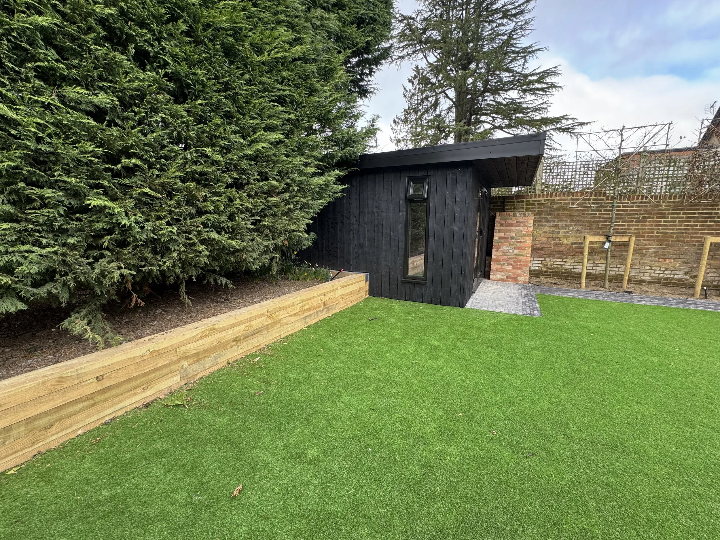 Small modern black wooden shed with tall narrow window, attached brick chimney, paved pathway, green artificial lawn, and large evergreen tree in the background.