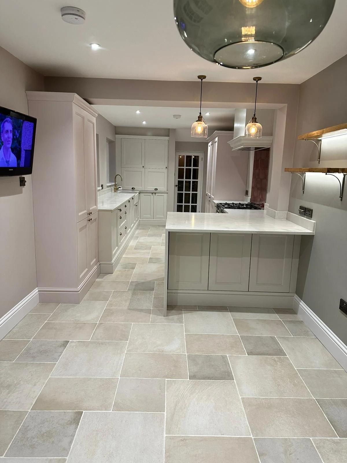 Modern kitchen with white cabinets, beige tiled floor, a small island, pendant lighting, and a wall-mounted TV.