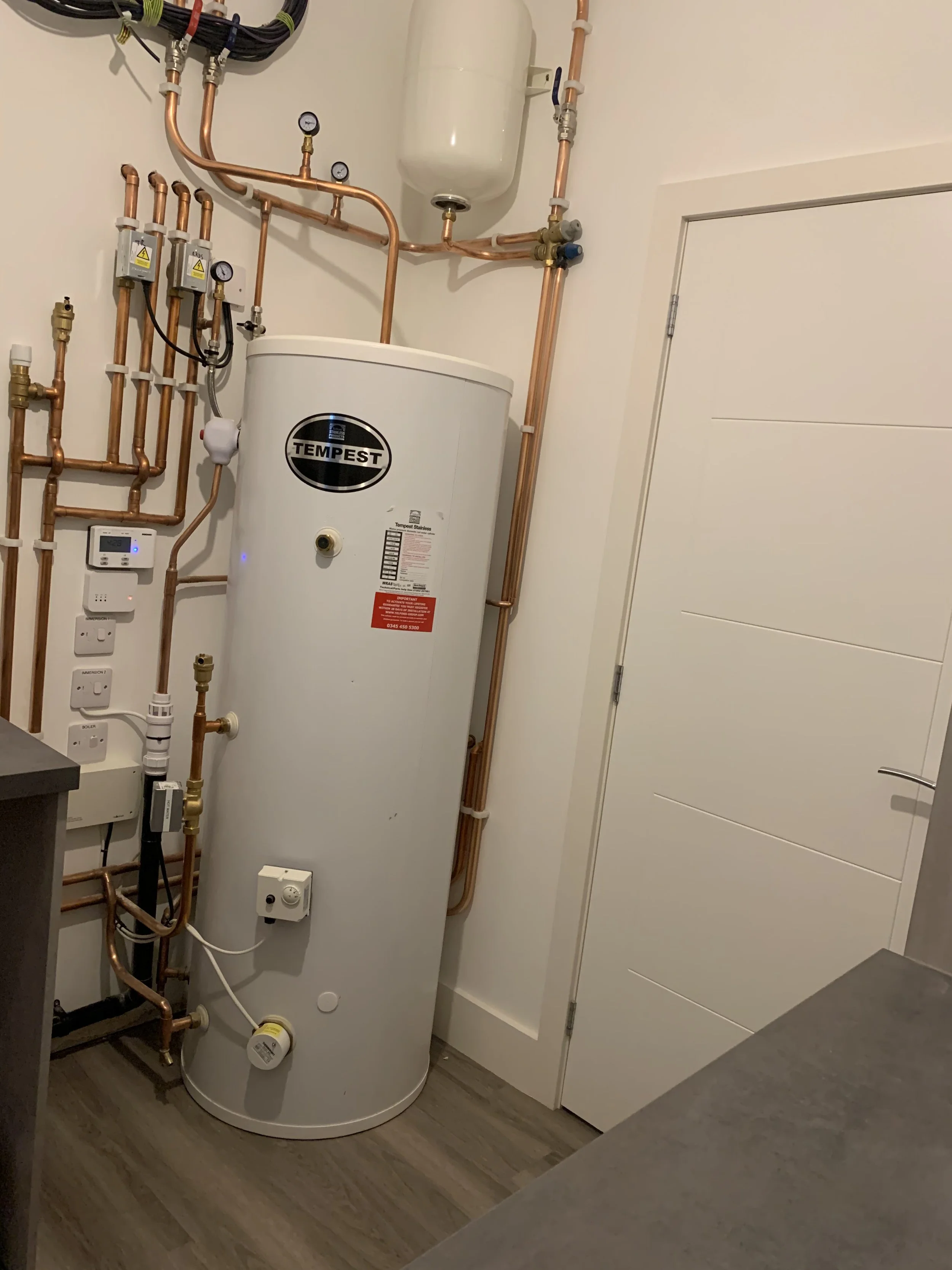 A hot water heater labeled 'Tempest' with copper pipes, pressure gauges, and control panels surrounding it in a utility room.
