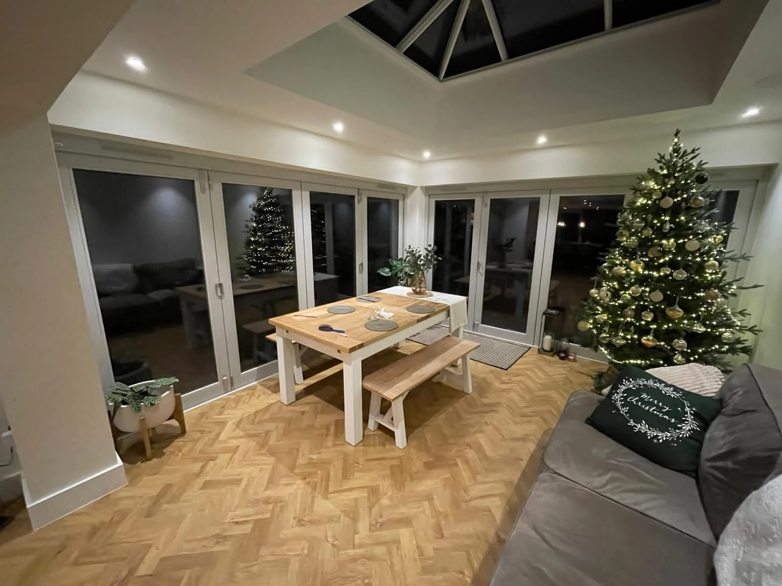 A living room decorated for Christmas with a large lit Christmas tree, a dining table with wood and white accents, potted plant, sofa with a holiday pillow, and large glass sliding doors leading outside.