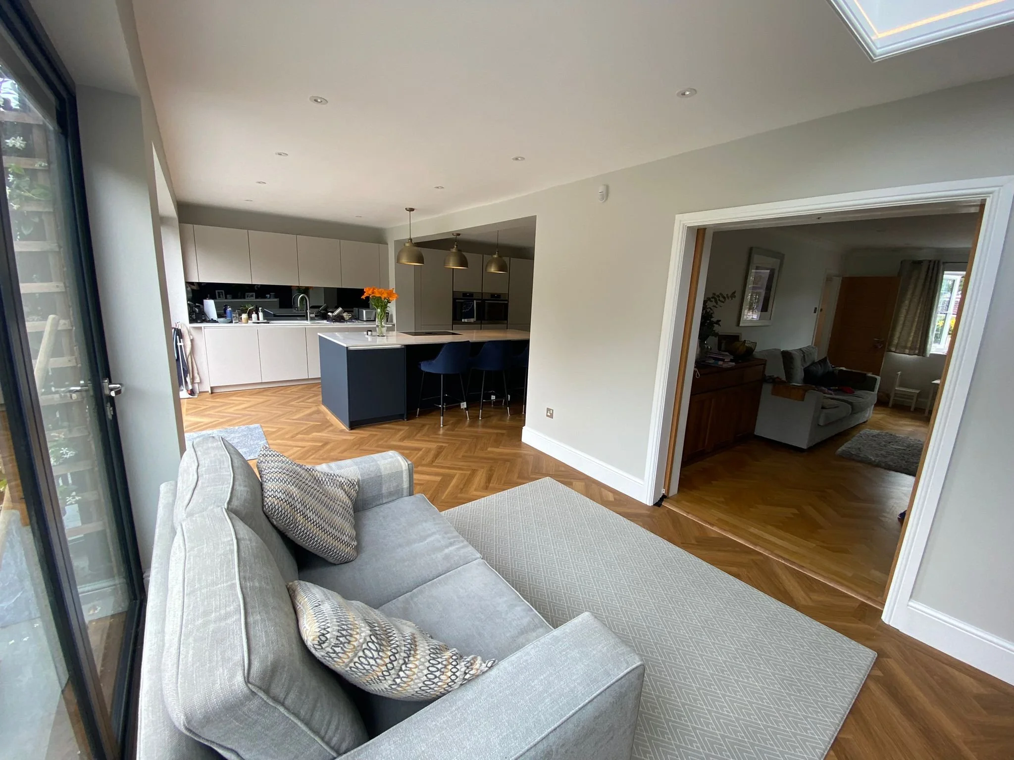 Open-concept living room and kitchen with hardwood floors, light-colored walls, and a skylight ceiling. A light gray sofa with patterned pillows is in the foreground, facing the kitchen area with a black island and white cabinets. Adjacent to the liv