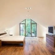 Spacious bedroom with a large window, wooden floor, and minimal furniture including a bed and a TV on a dresser.