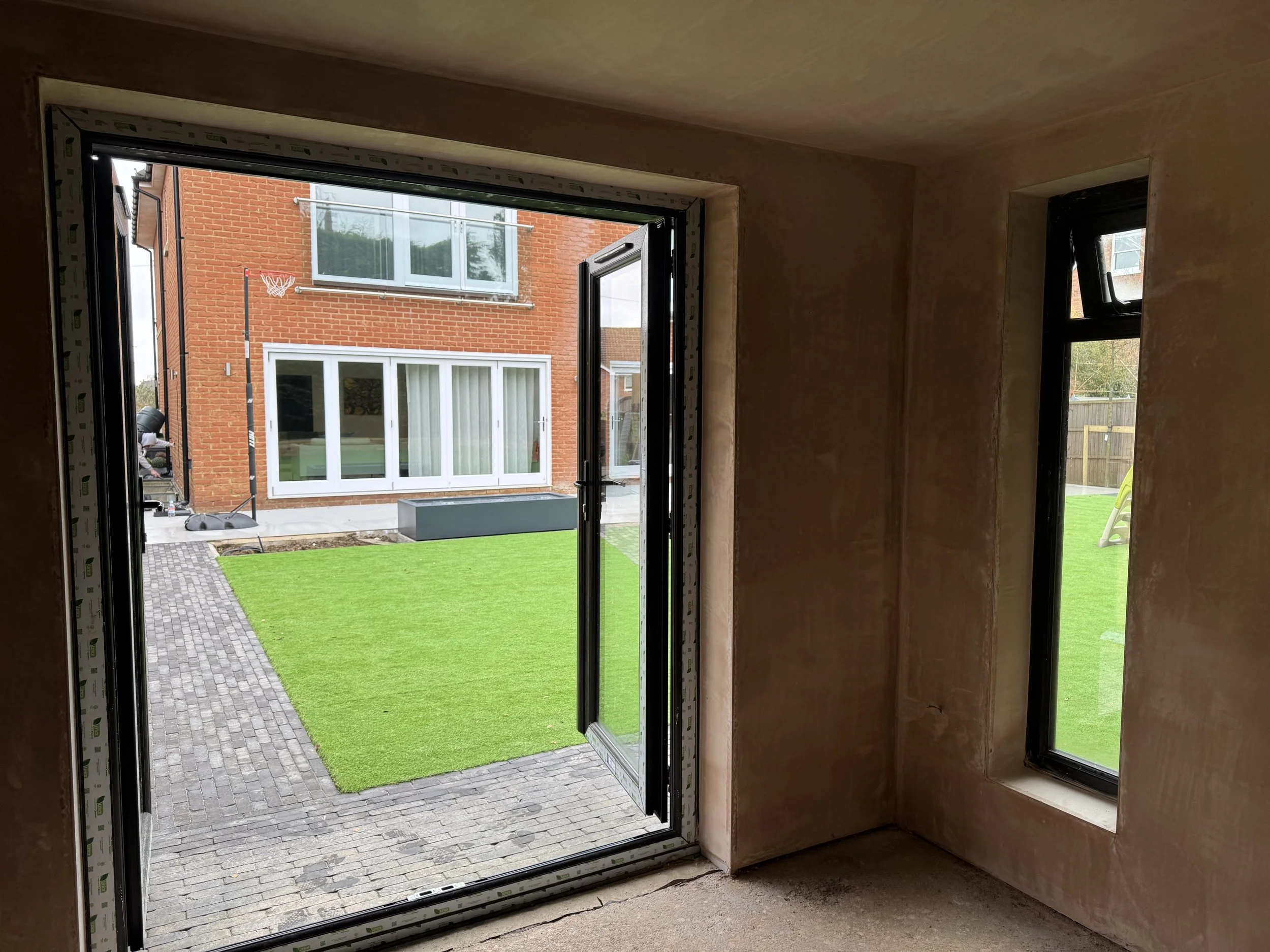 Interior view of a room under construction with a large open door leading to a backyard with green artificial turf and a brick house with large windows.
