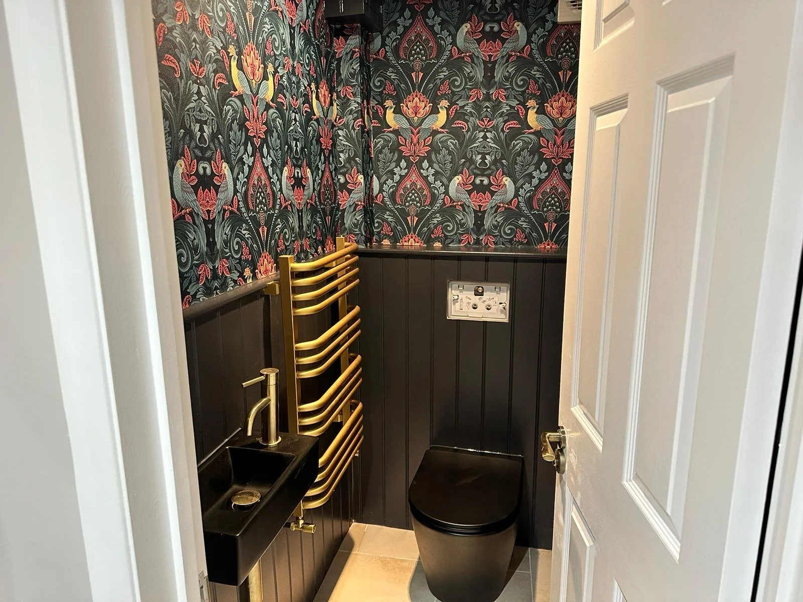 Small bathroom with black toilet, a black sink with a gold faucet, a folded gold towel rack, dark floral wallpaper with pink, green, and yellow accents, white door partially open, and beige floor tiles.
