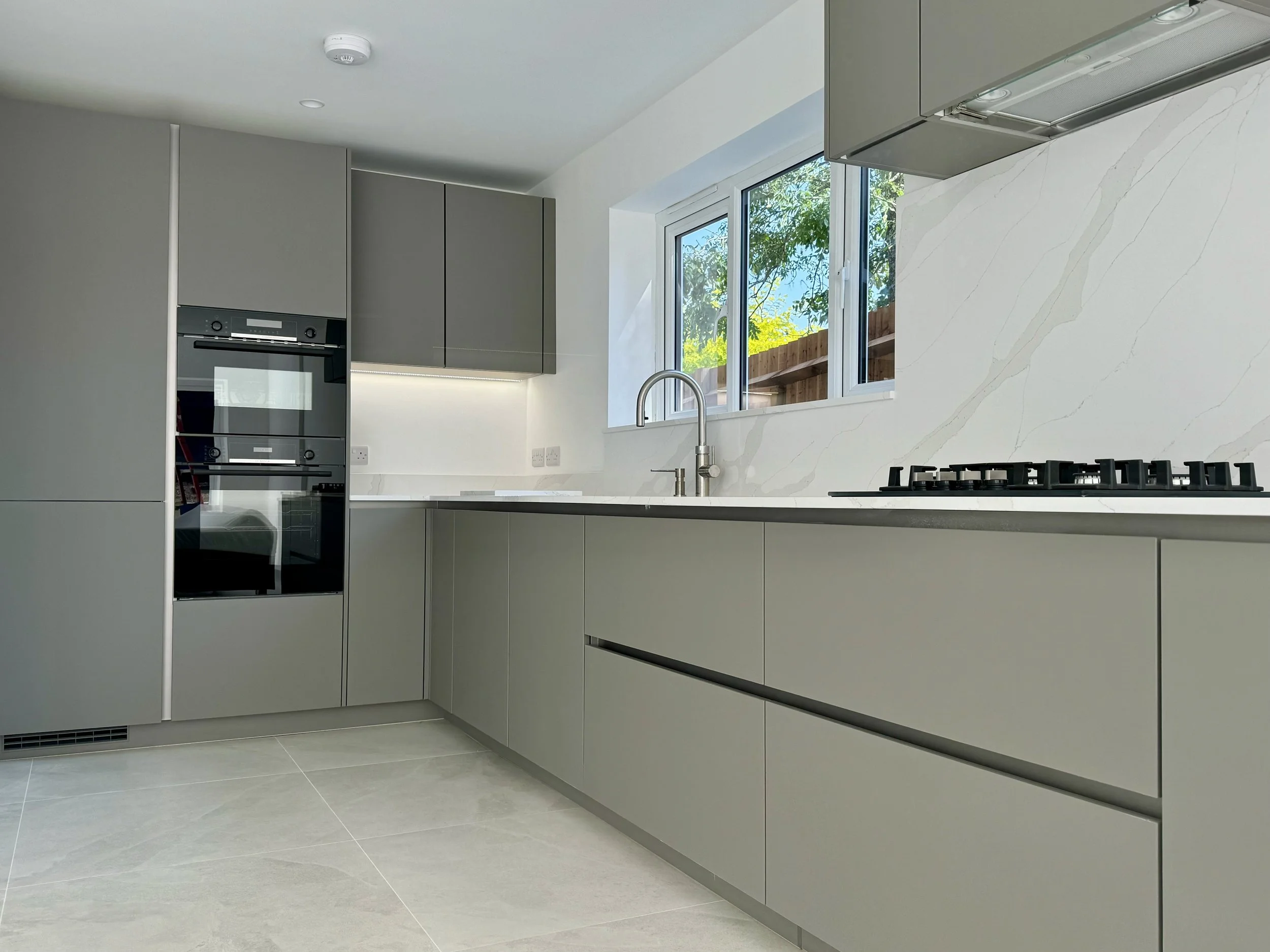 Modern kitchen with light gray cabinetry, a gas stove, a built-in oven, and a window showing outdoor trees and yard.
