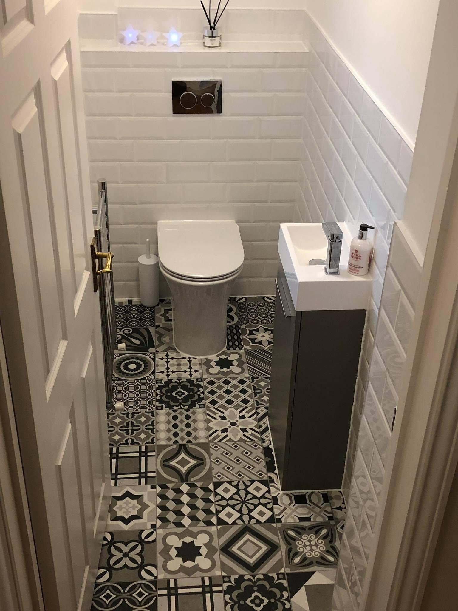 Small modern bathroom with patterned black, white, and gray tiles on the floor, a wall-mounted toilet with a black water flush panel, a small white sink with a gray cabinet, and decorative star-shaped lights on a shelf above the toilet.