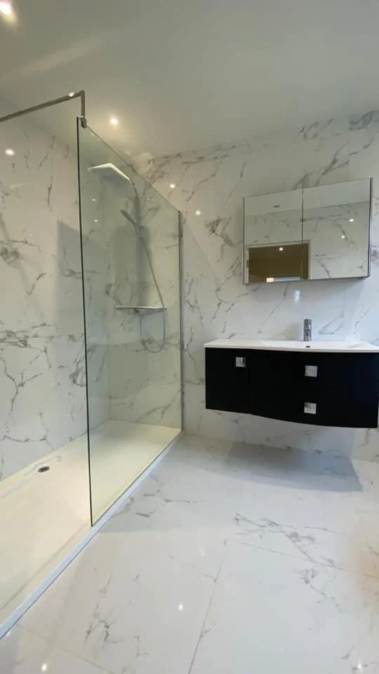 Modern bathroom with marble walls and floors, a glass-enclosed shower area with a rain showerhead, black vanity with a white countertop, and a mirrored cabinet above the sink.