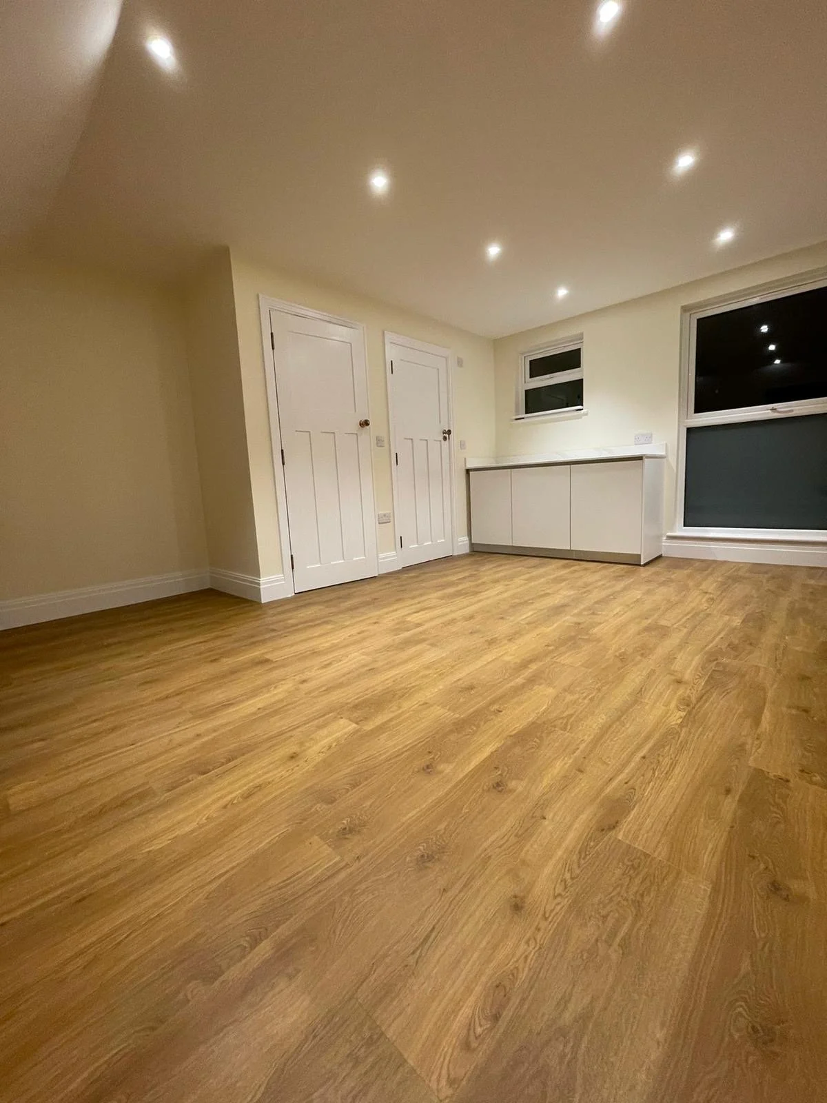 Empty room with wooden floor, white walls, ceiling with recessed lighting, two white doors, a window, and a built-in white cabinet.