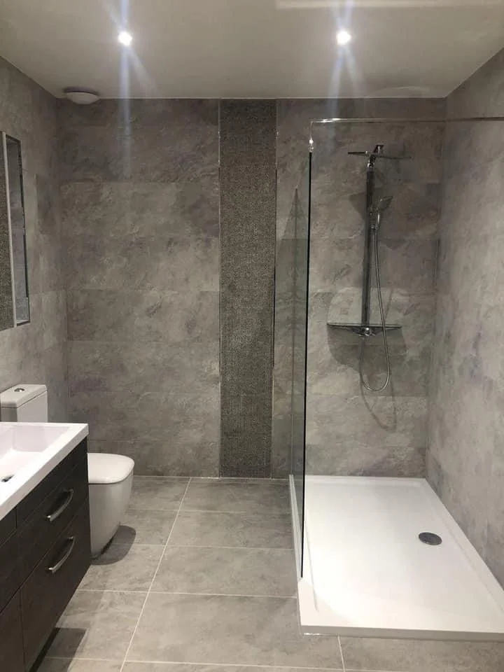 Modern bathroom with gray tiled walls and floor, a walk-in shower with glass partition, a white toilet, and a black vanity with a sink.
