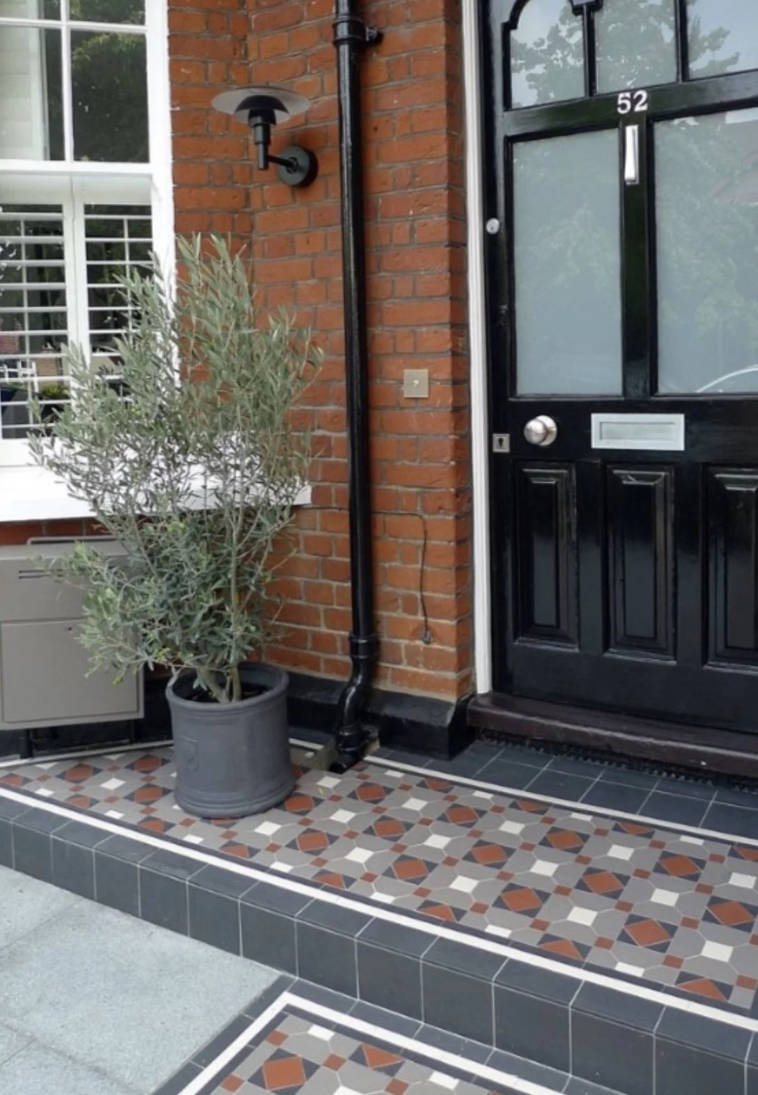 Front porch of a house featuring a black door with a brass handle and mail slot, a potted plant, a brick wall, a black drainpipe, and decorative patterned tiles on the porch floor.