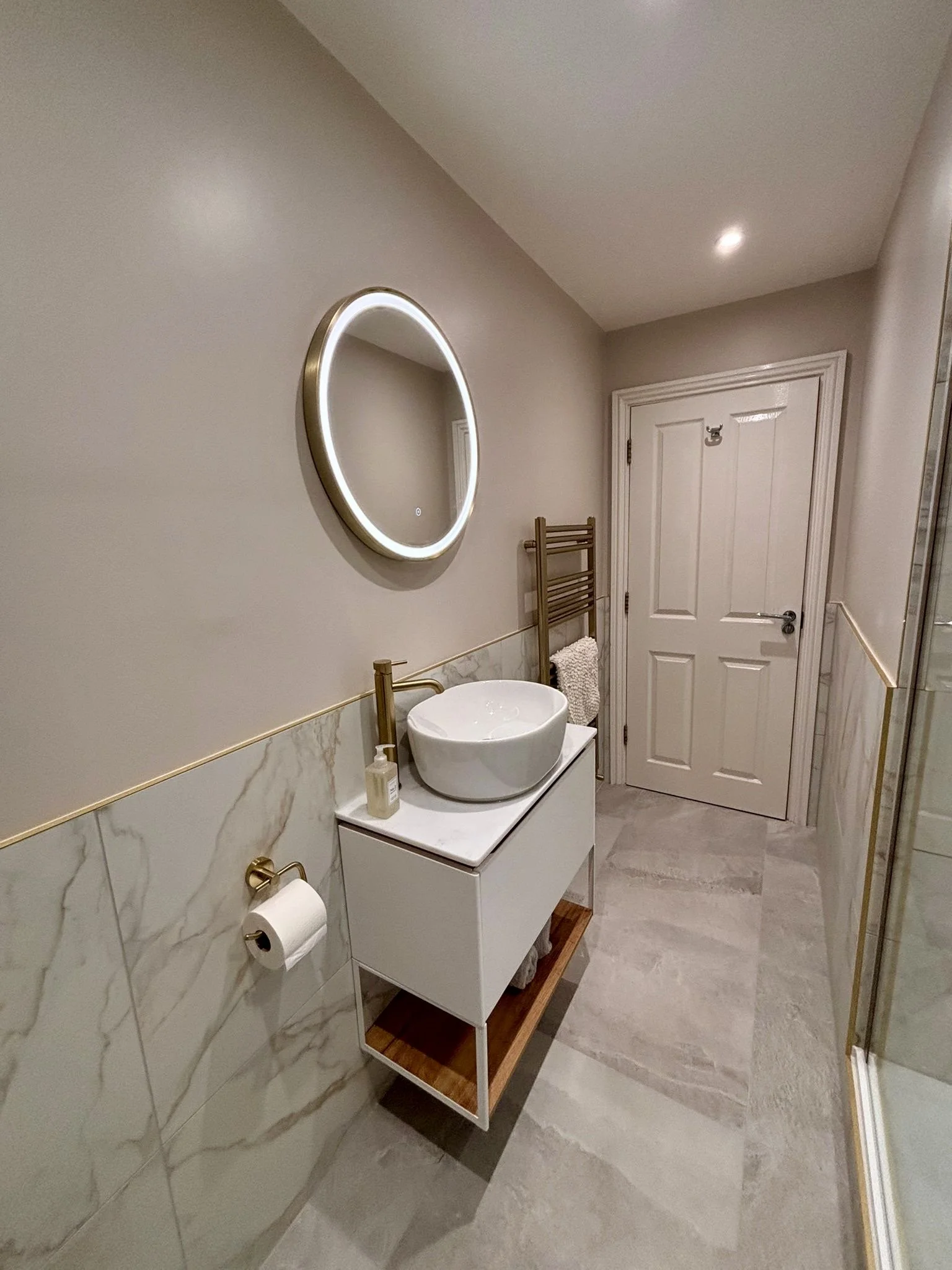 A modern bathroom with a circular mirror with LED lighting, a white vessel sink on a sleek vanity with a wooden shelf, a towel rack, a bottle of soap, a roll of toilet paper, a closed white door, and beige marble tiles on the lower half of the wall, 