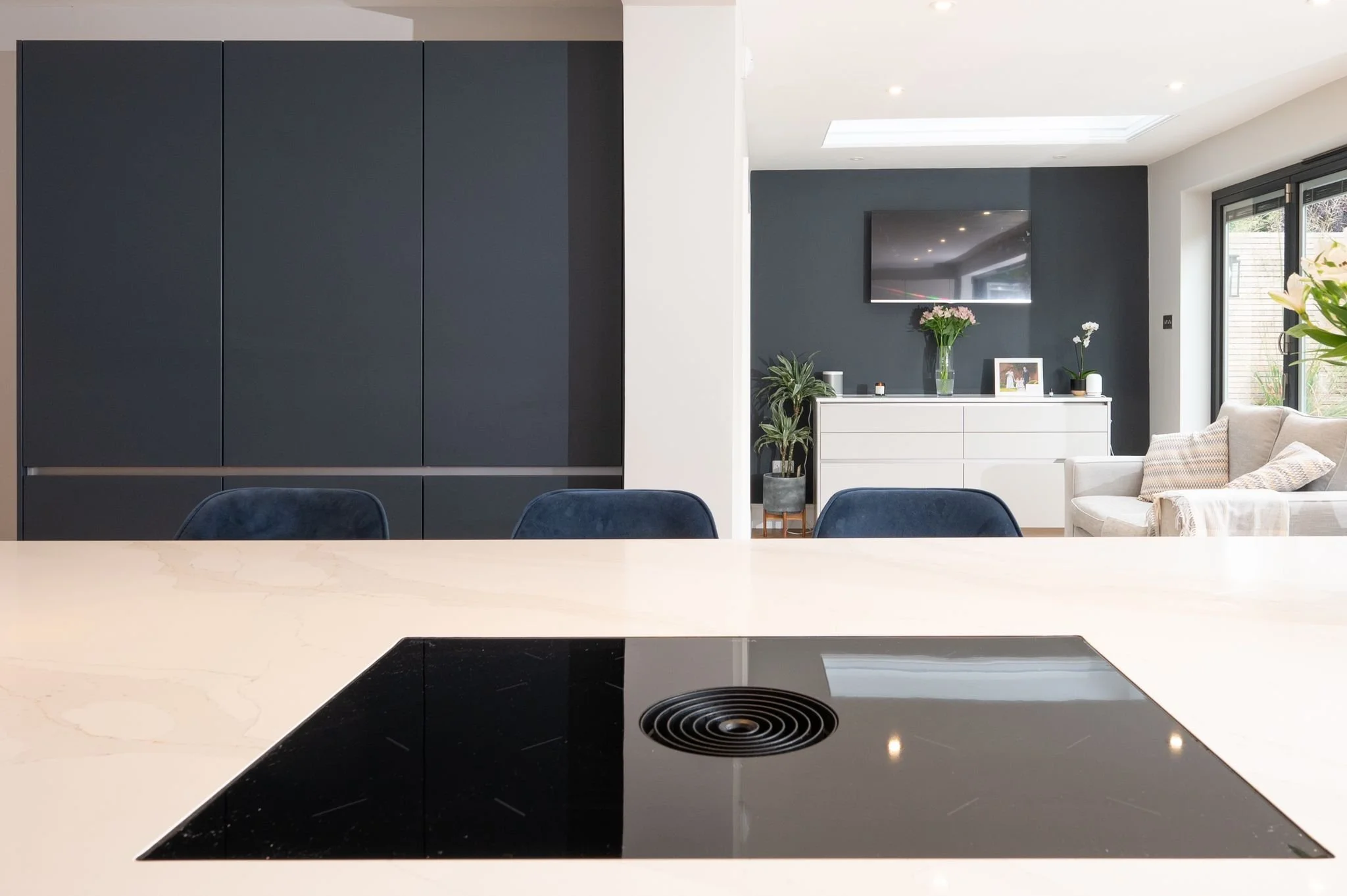 Modern kitchen with white island counter, black cooktop, navy blue chairs, dark blue wall cabinets, and a living area with a white sideboard, a flat-screen TV, potted plants, and a beige sofa near large windows with natural light.