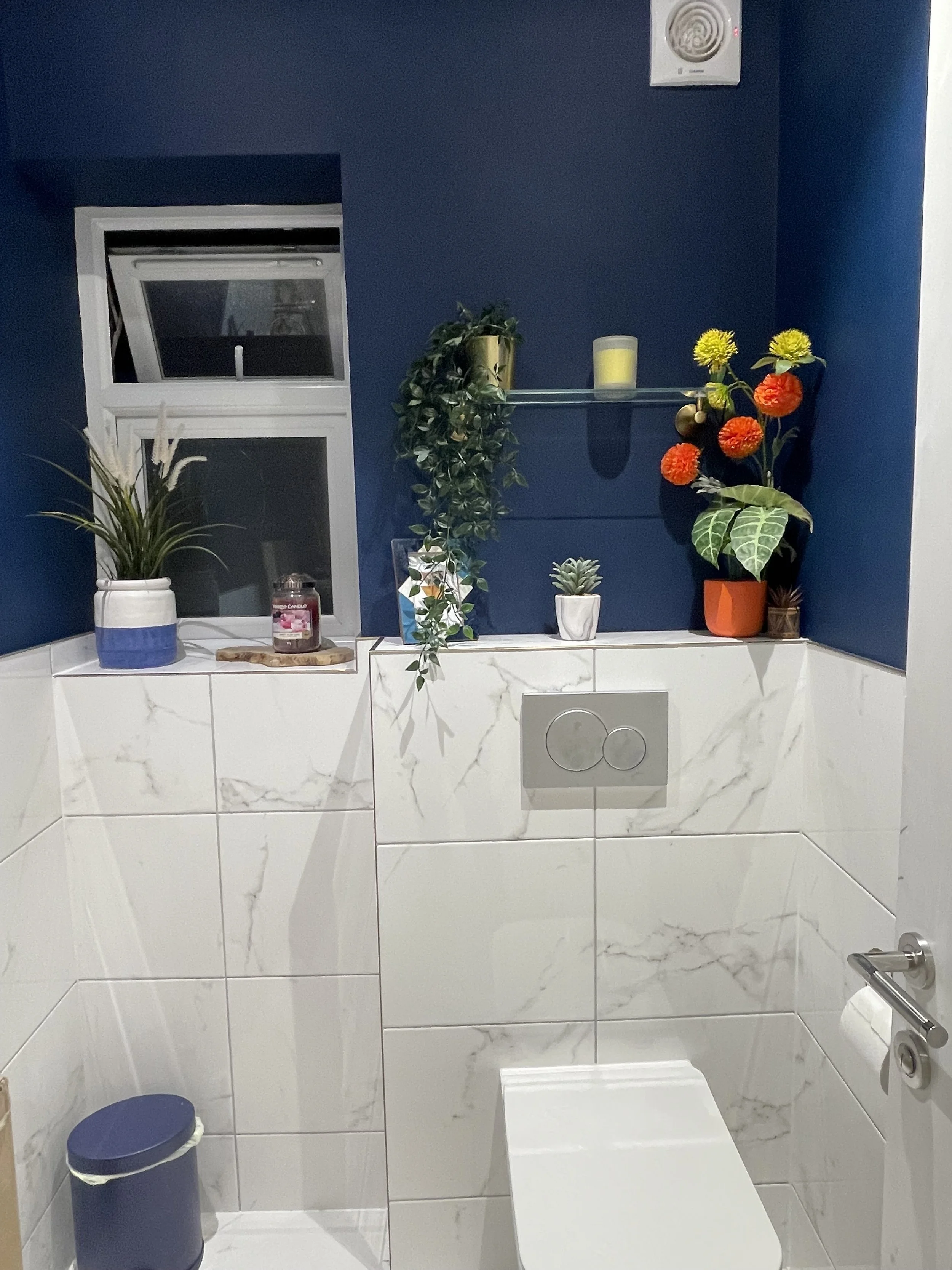 A modern bathroom with white marble tiles, a blue wall, and a small window. Decor includes various potted plants on a ledge and shelf, along with a candle and decorative items. A toilet is visible below the shelf, with a blue trash can nearby.