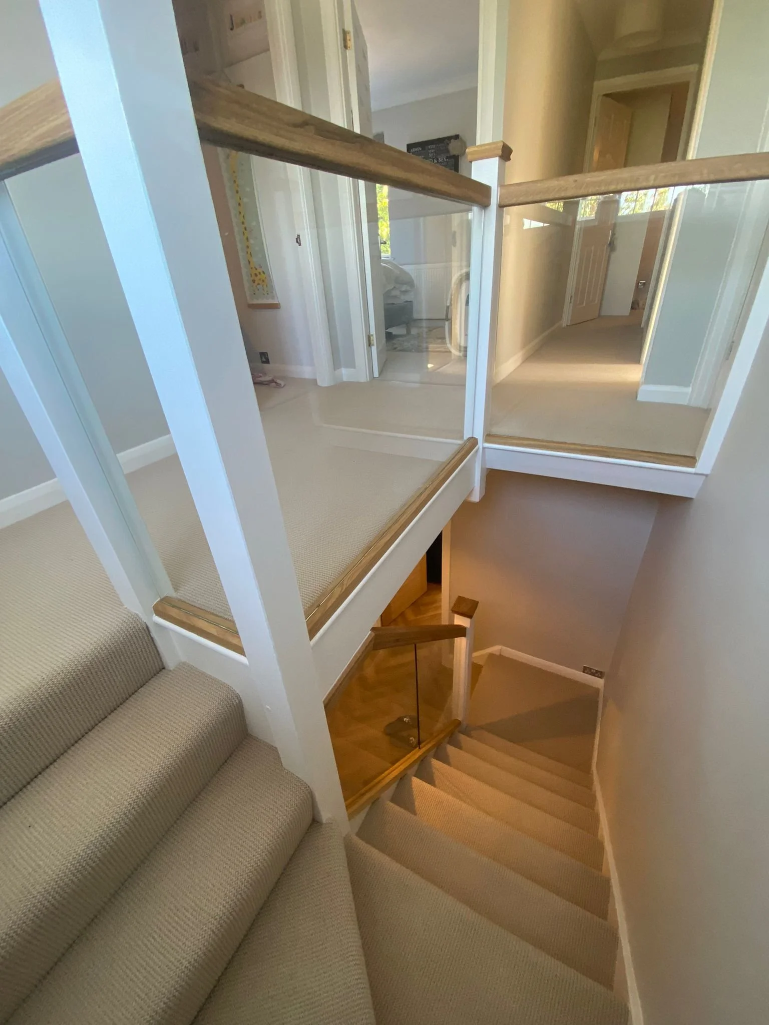 A staircase with beige carpet and a glass-paneled wooden handrail, leading down to a hallway with beige walls and doors.