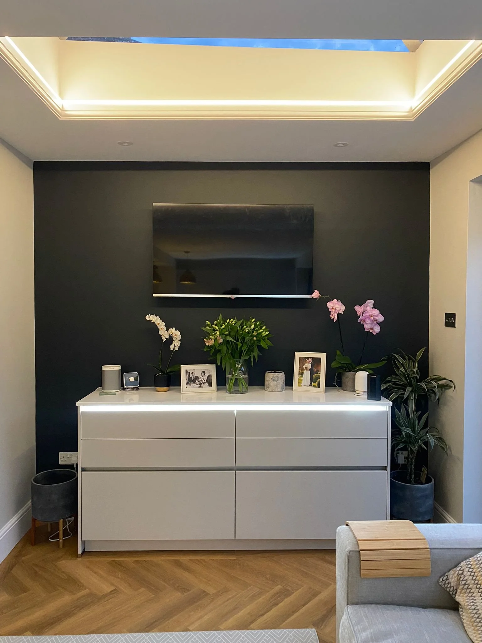 Modern living room with a white sideboard decorated with potted plants, framed photos, and small speakers, against a dark accent wall and a ceiling with ambient lighting.