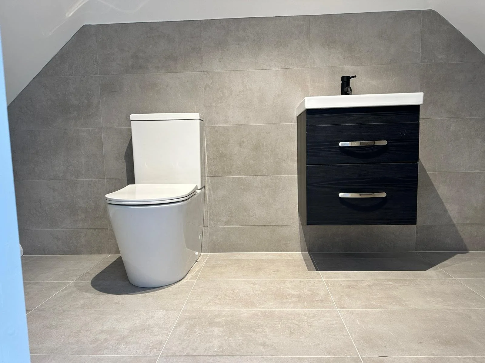 Modern bathroom with a white toilet and a black floating vanity with a white sink and black faucet, against gray tiled walls and floor.