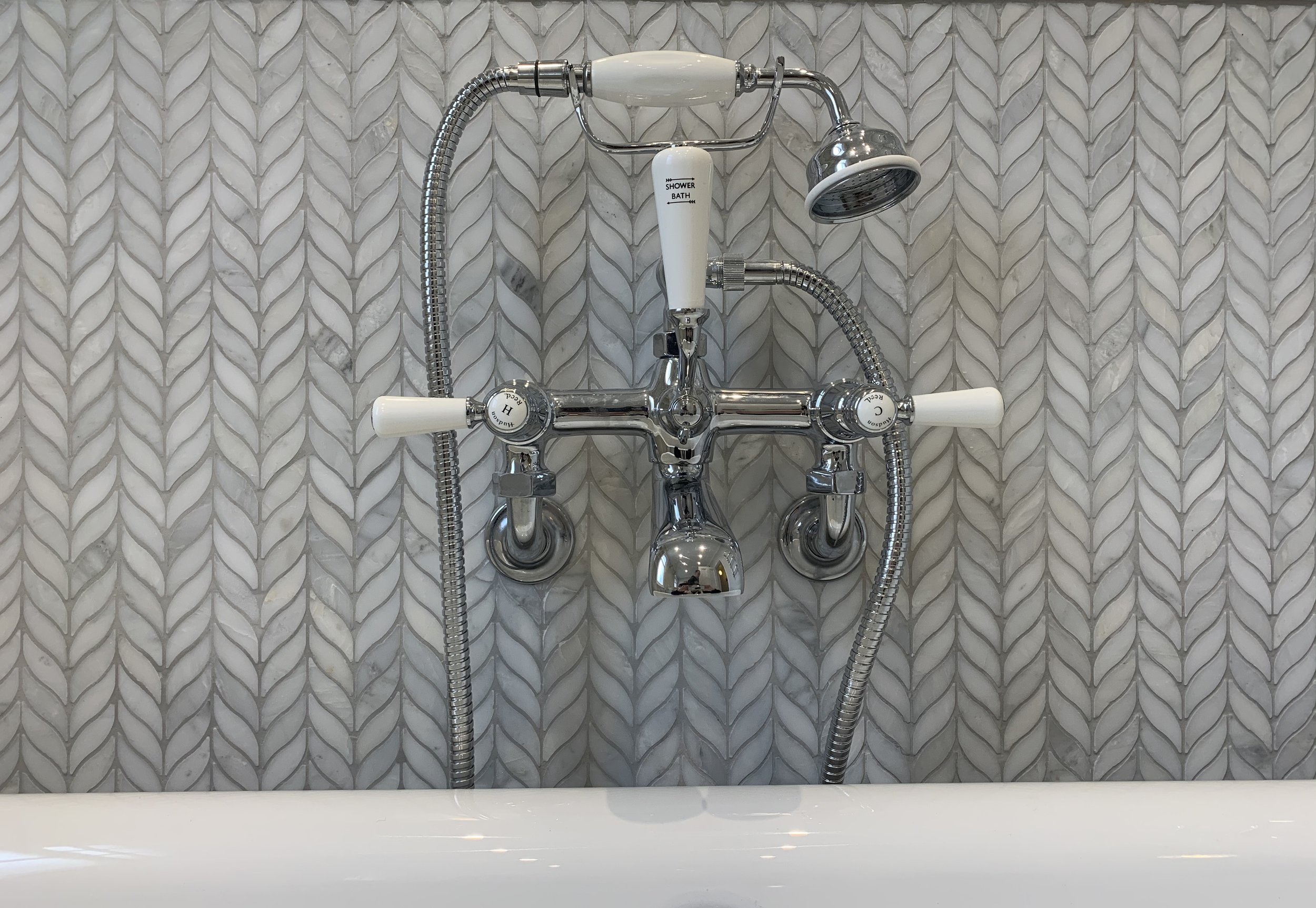 A silver bathtub faucet with hot and cold handles, a shower head, and flexible hoses mounted on a wall with gray tiles that have a leaf-like pattern.