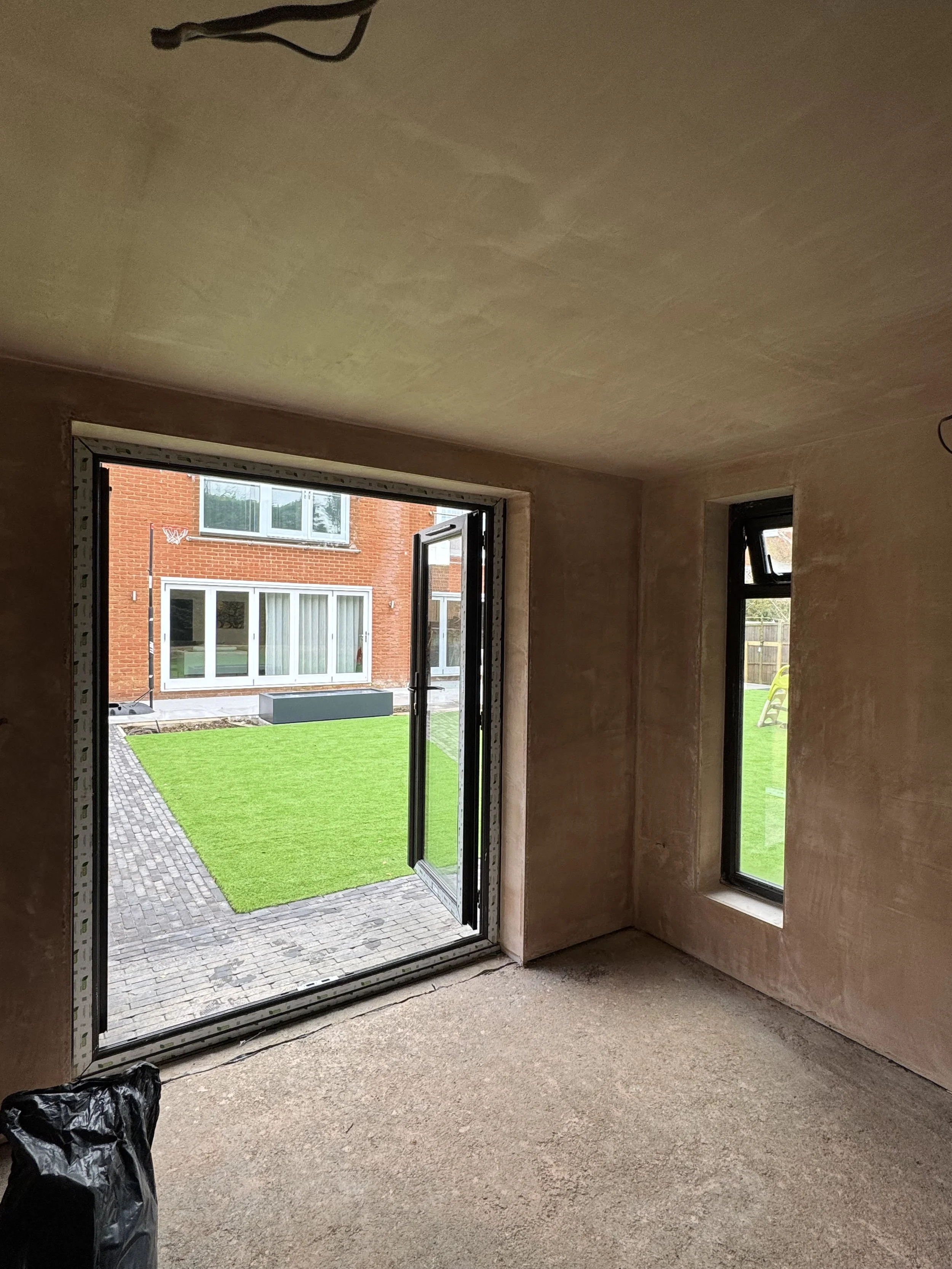 Interior view of a room under construction with a large glass door open to an outdoor garden with artificial grass and a neighboring brick house.