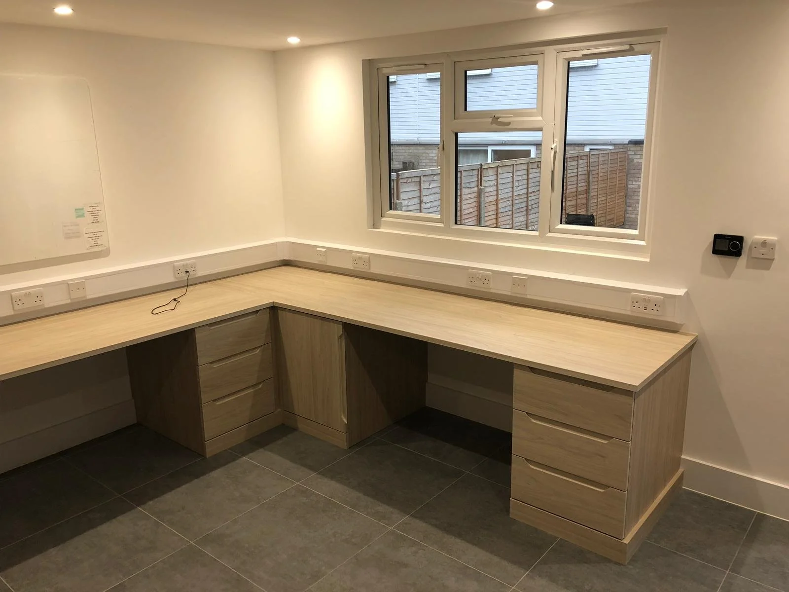 Empty office corner with wooden desk, multiple power outlets, white walls, and a window showing a fenced outdoor area.