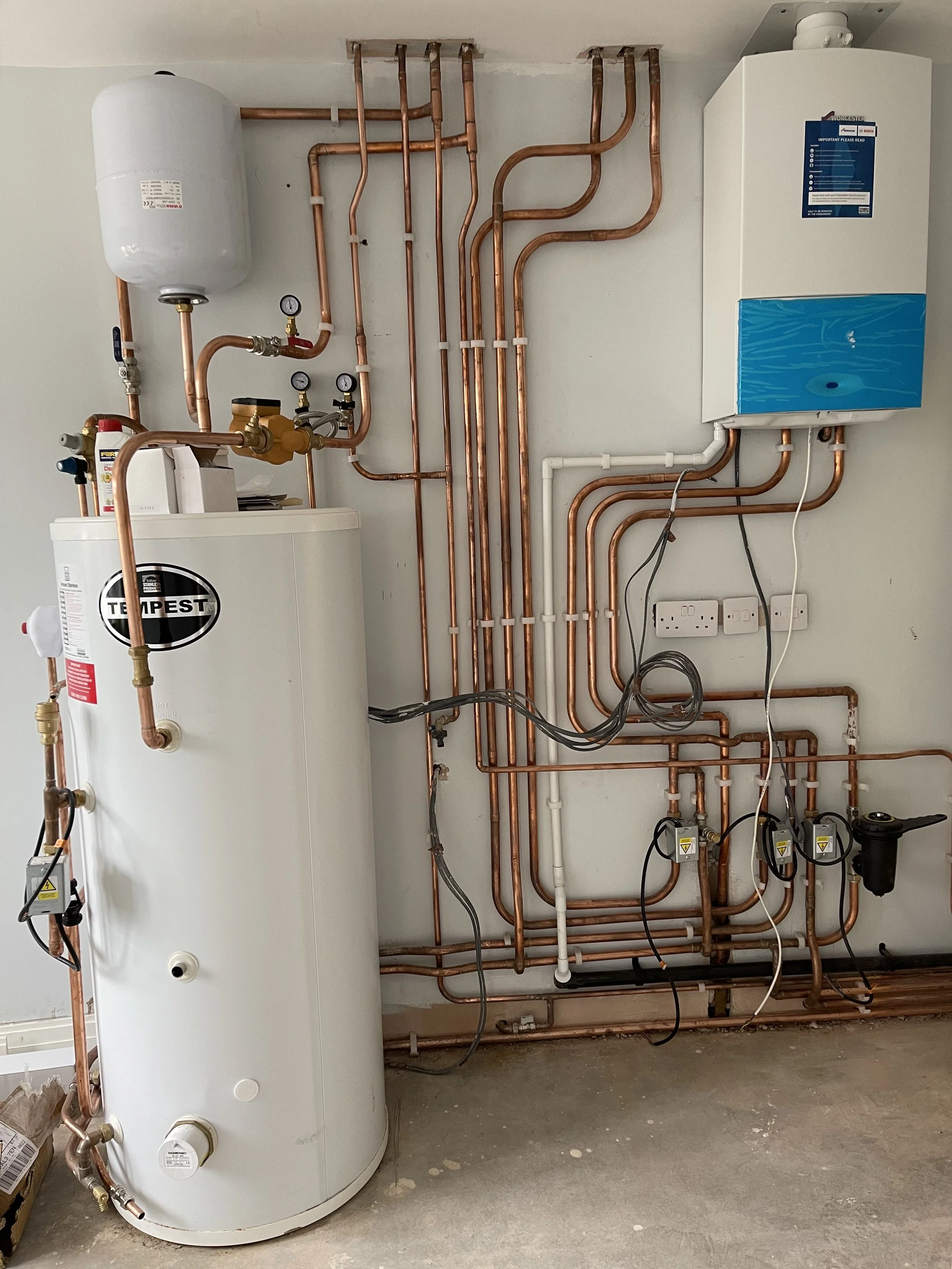 Installation of a domestic water heating system with copper pipes, a white hot water tank, and a wall-mounted boiler.