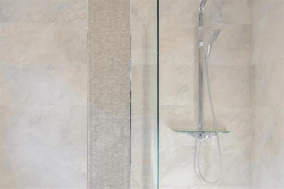 Shower area with beige tile walls, a glass partition, a handheld showerhead, and a small glass shelf.