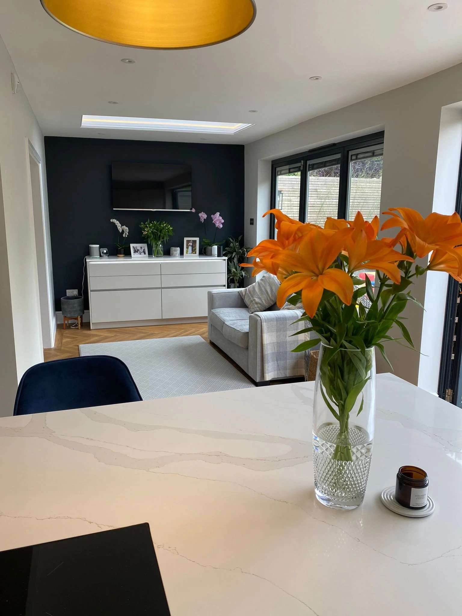 A modern living room with a white marble table, orange lilies in a glass vase, and a TV on a dark accent wall. There is a window with sliding doors, a gray sofa with cushions, and a white cabinet with pictures and plants.