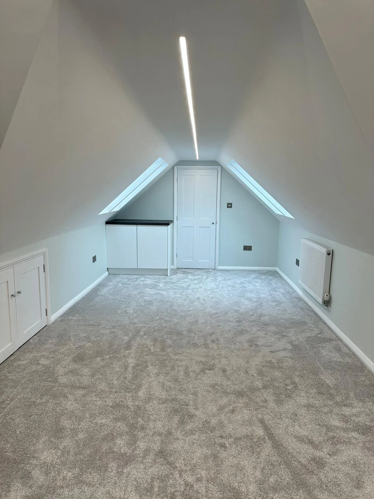 Empty attic room with beige carpet, white walls, two skylights, a white door, a small white cabinet, white radiators, and gray electrical outlets.