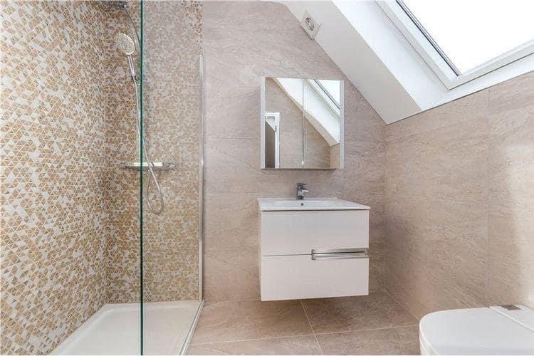 Bathroom with a walk-in shower on the left, a white vanity with a mirror above, a sloped ceiling with skylight, and beige tiled walls and floor.
