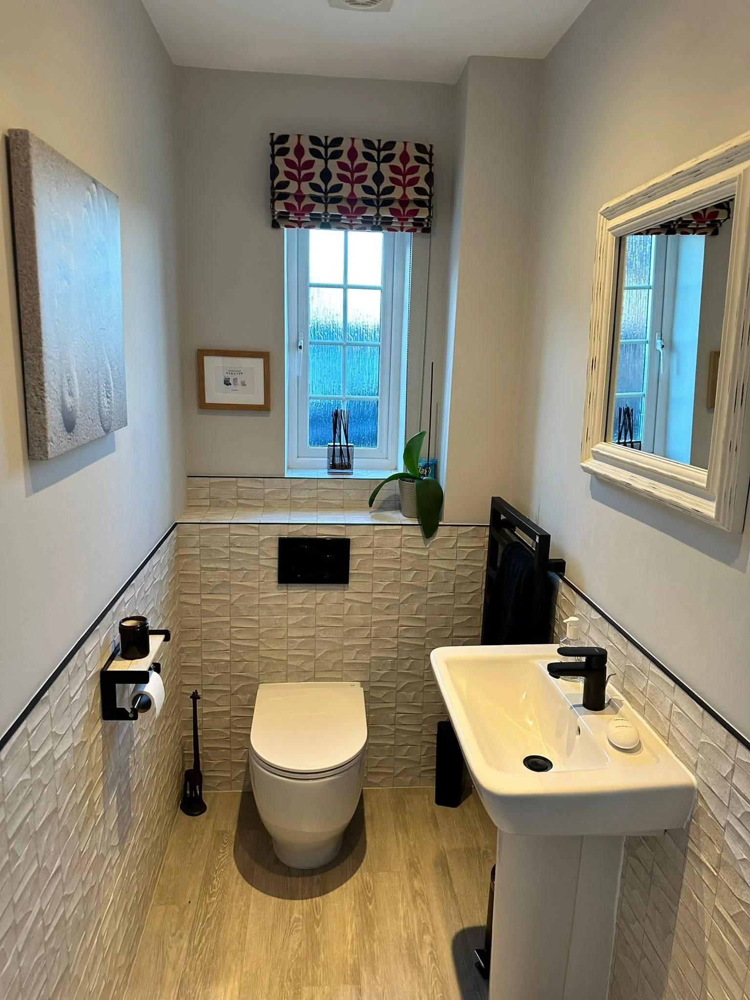 Small bathroom with a toilet, white sink, and a mirror, window with a valance, and a potted plant, decorated in neutral tones and minimalist style.