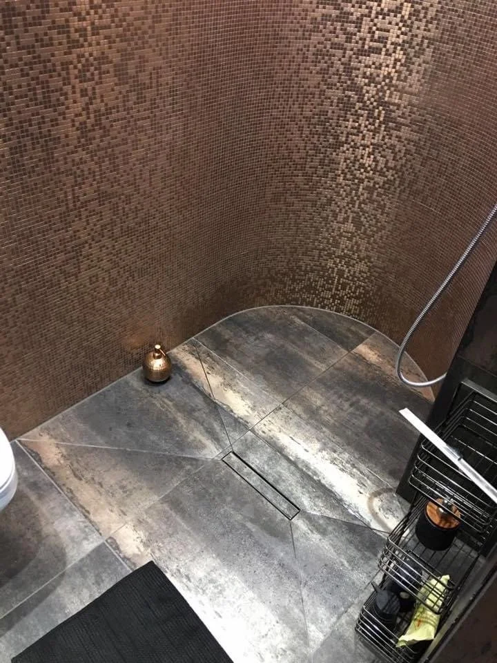 Corner of a bathroom shower area with brown mosaic tile wall, gray stone floor with drain, and a metallic soap dispenser on the floor. A black rack with toiletries and a yellow towel is visible.