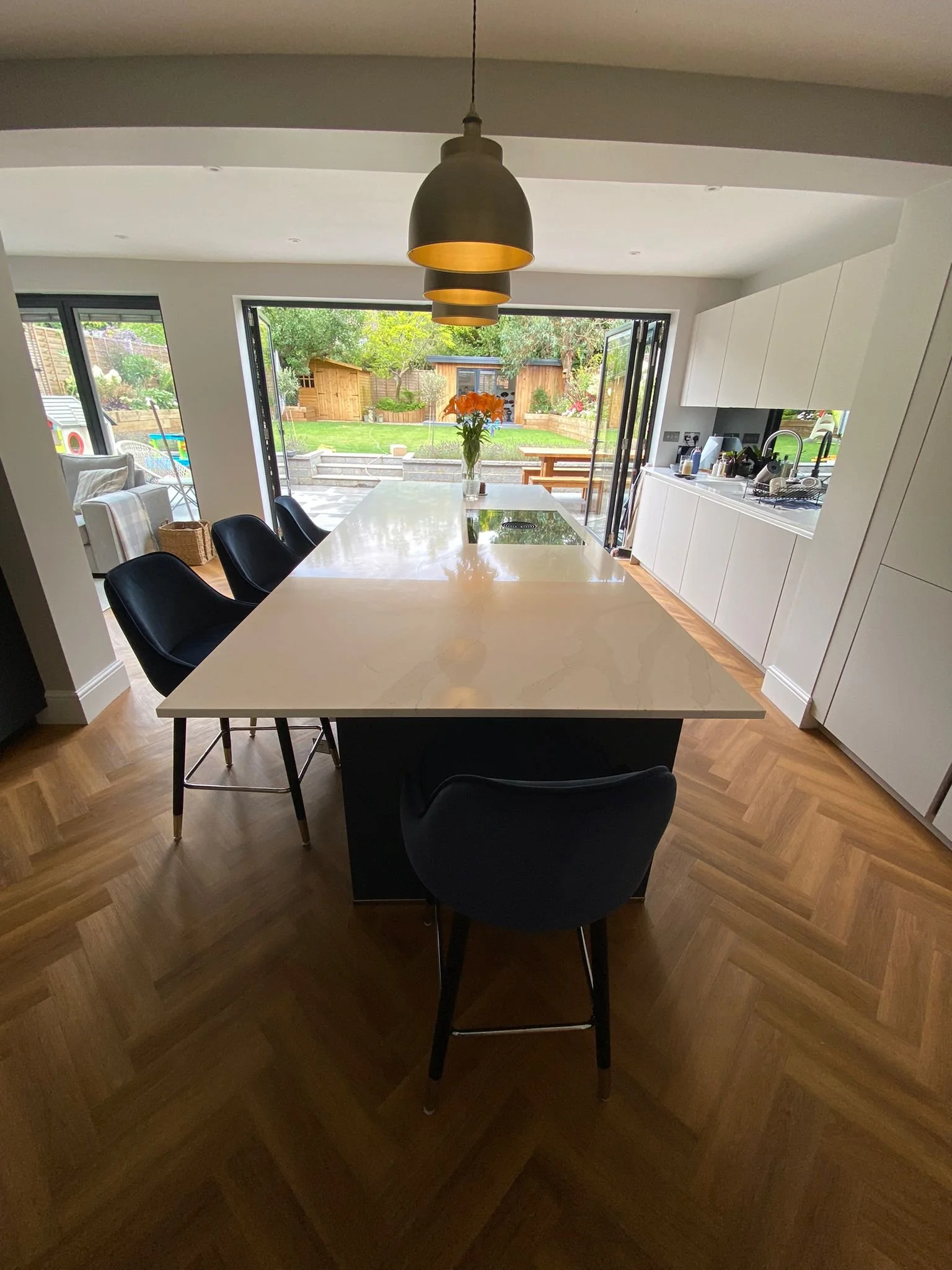 Modern kitchen with white countertops, black chairs around a kitchen island, open to a backyard with garden and wooden shed.