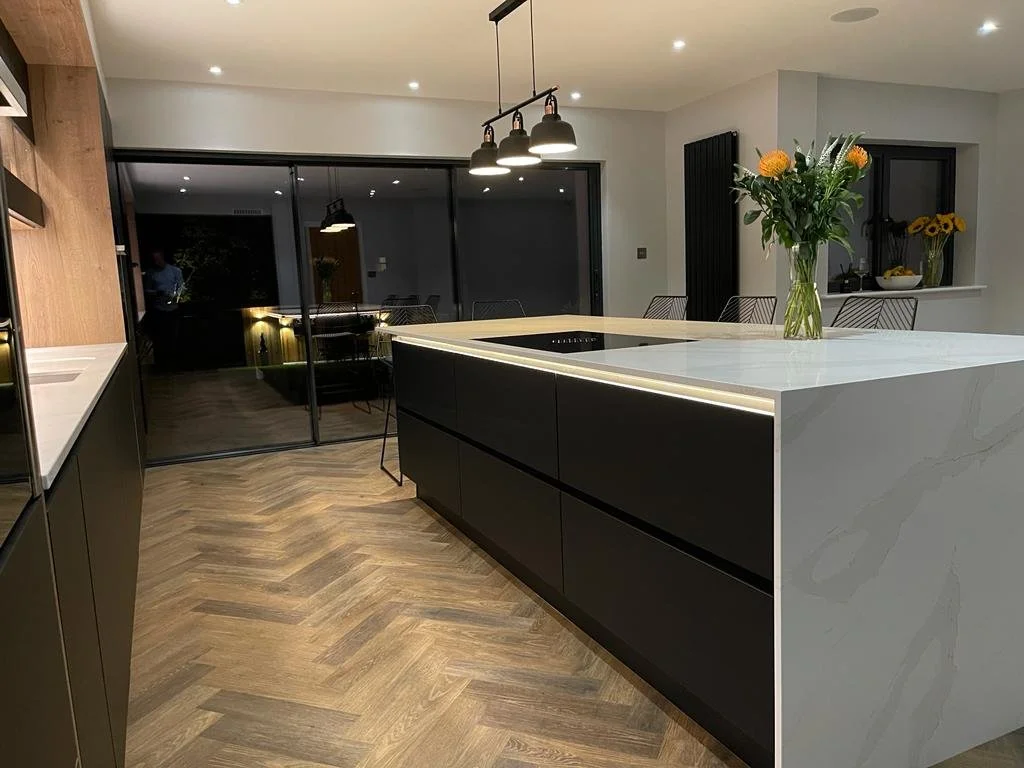 Modern kitchen with a large white marble island, black cabinetry, a vase of orange and yellow flowers, and a lighting fixture hanging above the island, with sliding glass doors and dining area in the background.