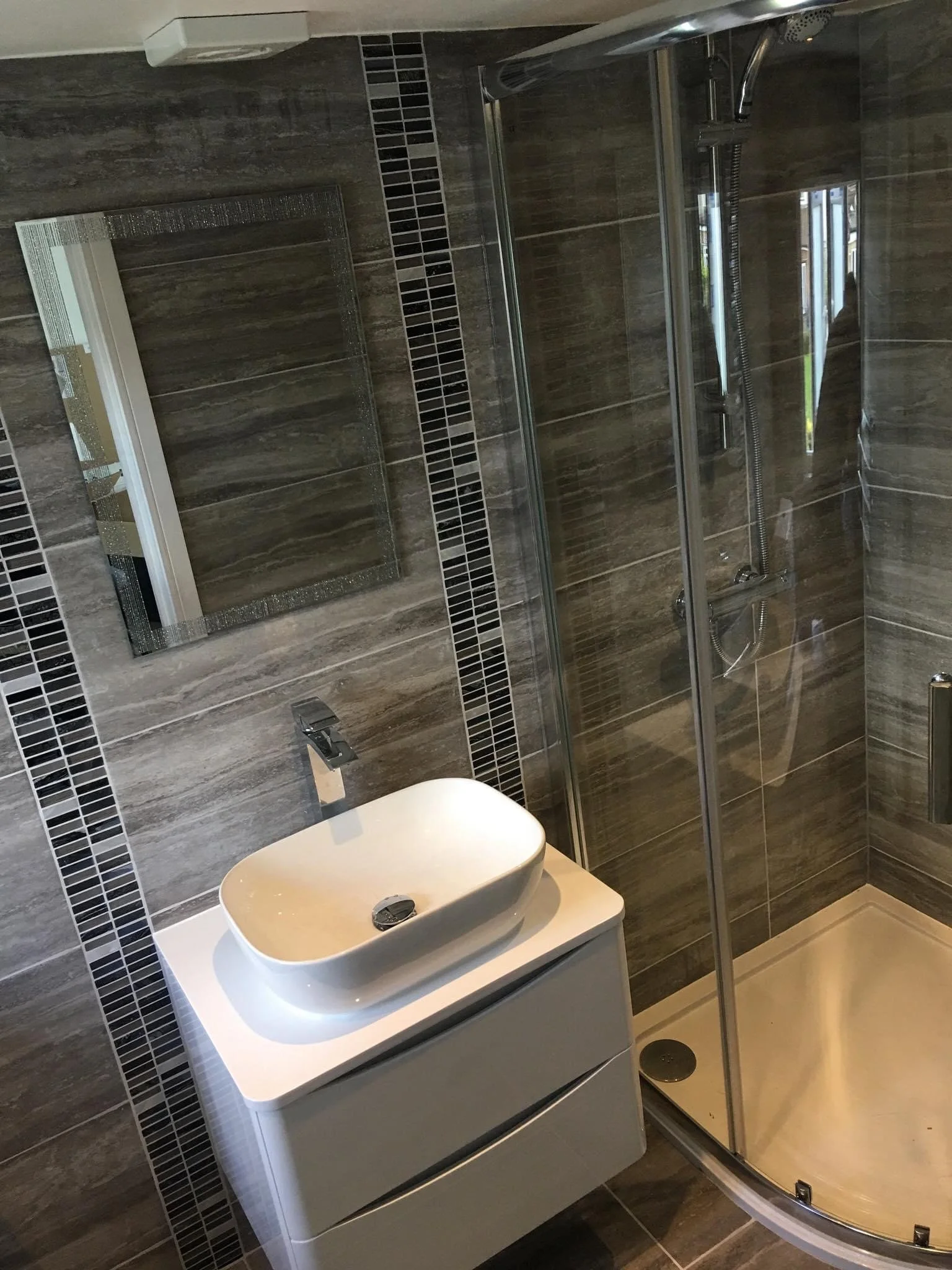 Modern bathroom with gray tiled walls, a rectangular mirror, a white vessel sink on a cabinet, and a glass-enclosed shower with a rainfall showerhead.