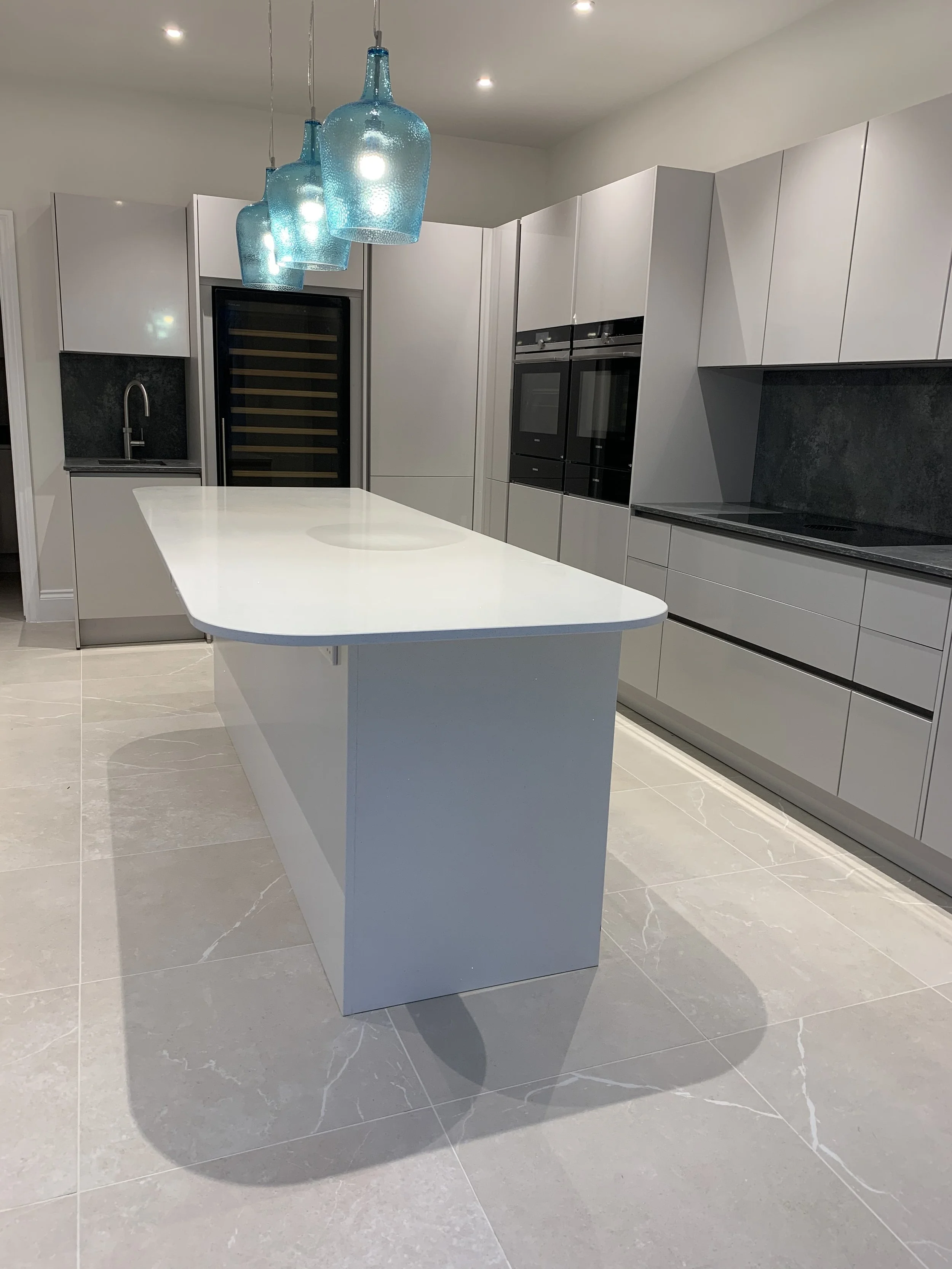 Modern kitchen with white cabinets, black appliances, a white island, gray tiled floor, and three blue glass pendant lights hanging from the ceiling.