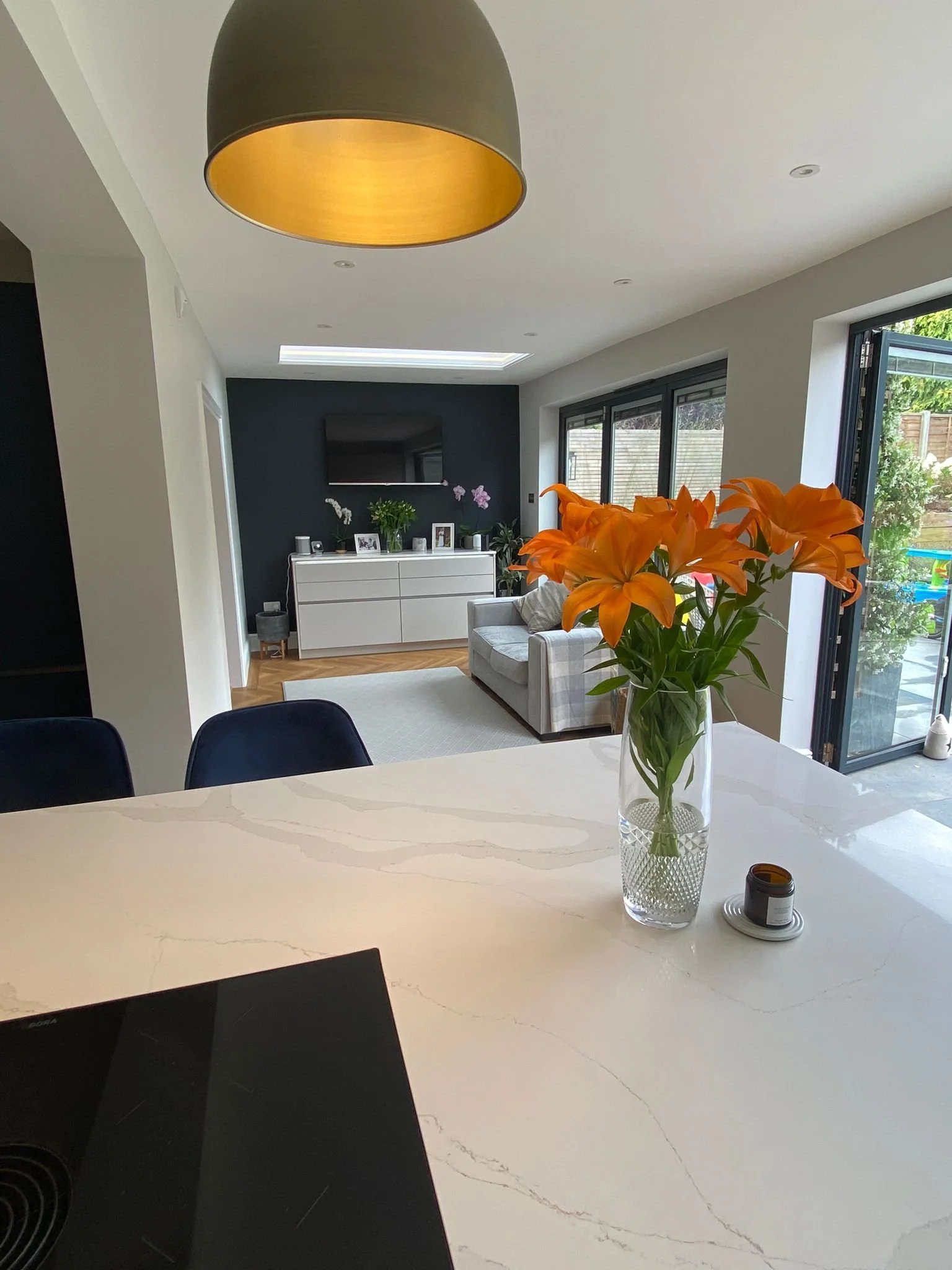 A modern, open-concept living room and kitchen area with a white marble kitchen island, orange lilies in a glass vase, and a glimpse of a gray seating area with a white TV console and black accent wall.