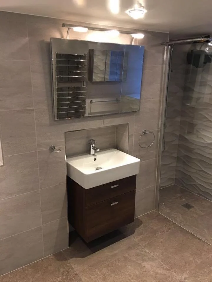 Bathroom with wall-mounted mirror, small sink with dark wooden cabinet, towel ring, and glass shower enclosure with textured tile walls.