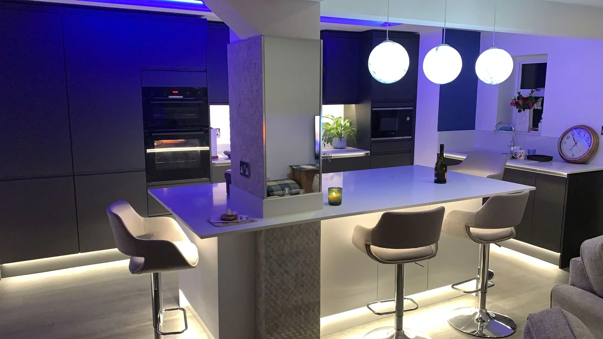 Modern kitchen with white island, three pendant lights, black cabinets, and a small window with flowers. There are bar stools at the island, and a lamp and clock on the countertop.