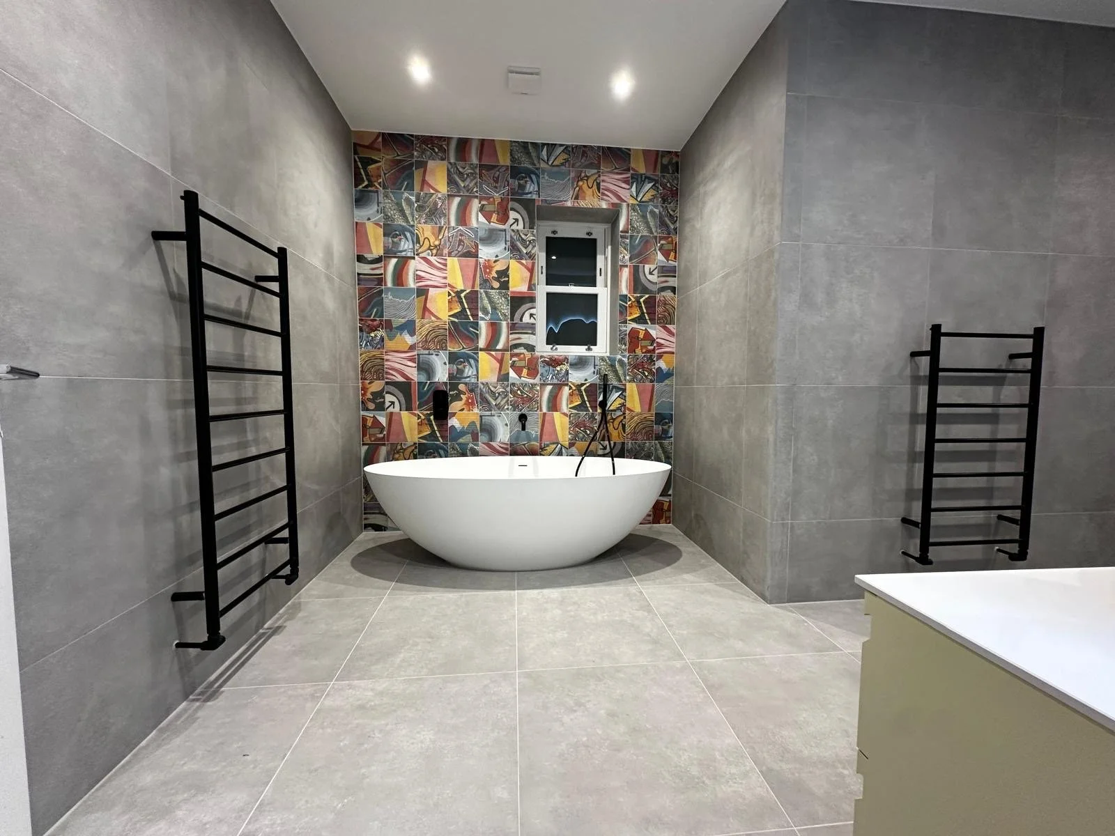 Modern bathroom with gray tiled walls and floor, a white oval bathtub, black heated towel racks, a colorful mosaic tile accent wall, and a small window.