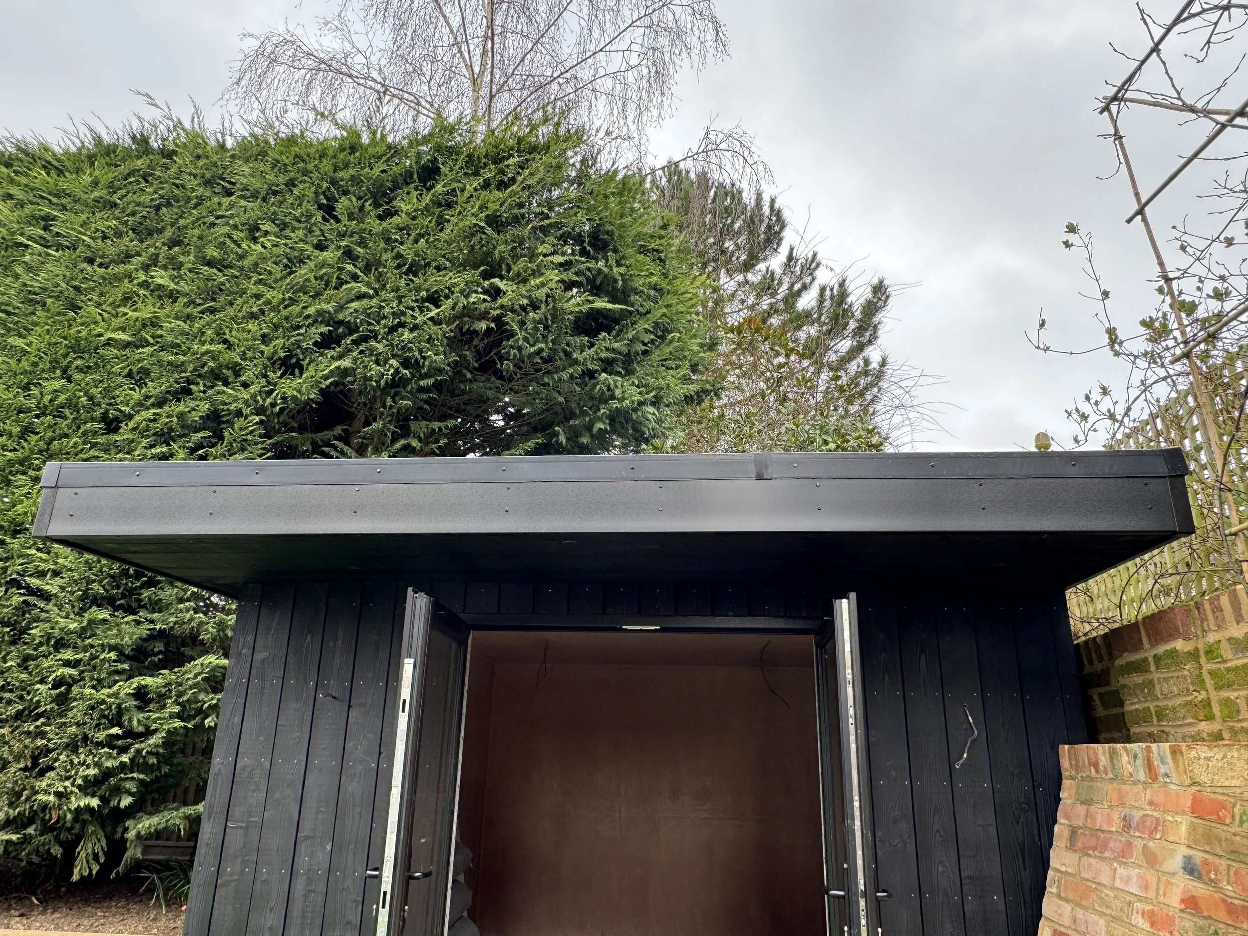 Dark-colored shed with open doors, surrounded by green trees and a brick wall, under a cloudy sky.