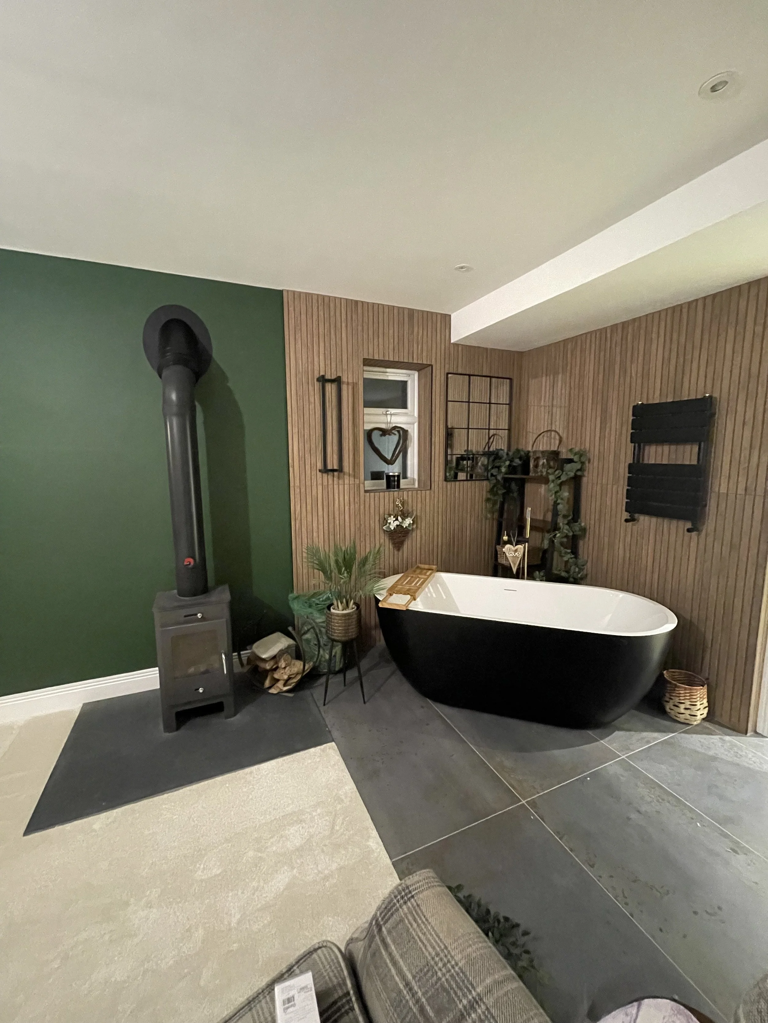 Indoor bathroom featuring a black bathtub with a wooden tray, wood-paneled wall with decorative items, green and black plants, a window, and a wood stove on a black rug.