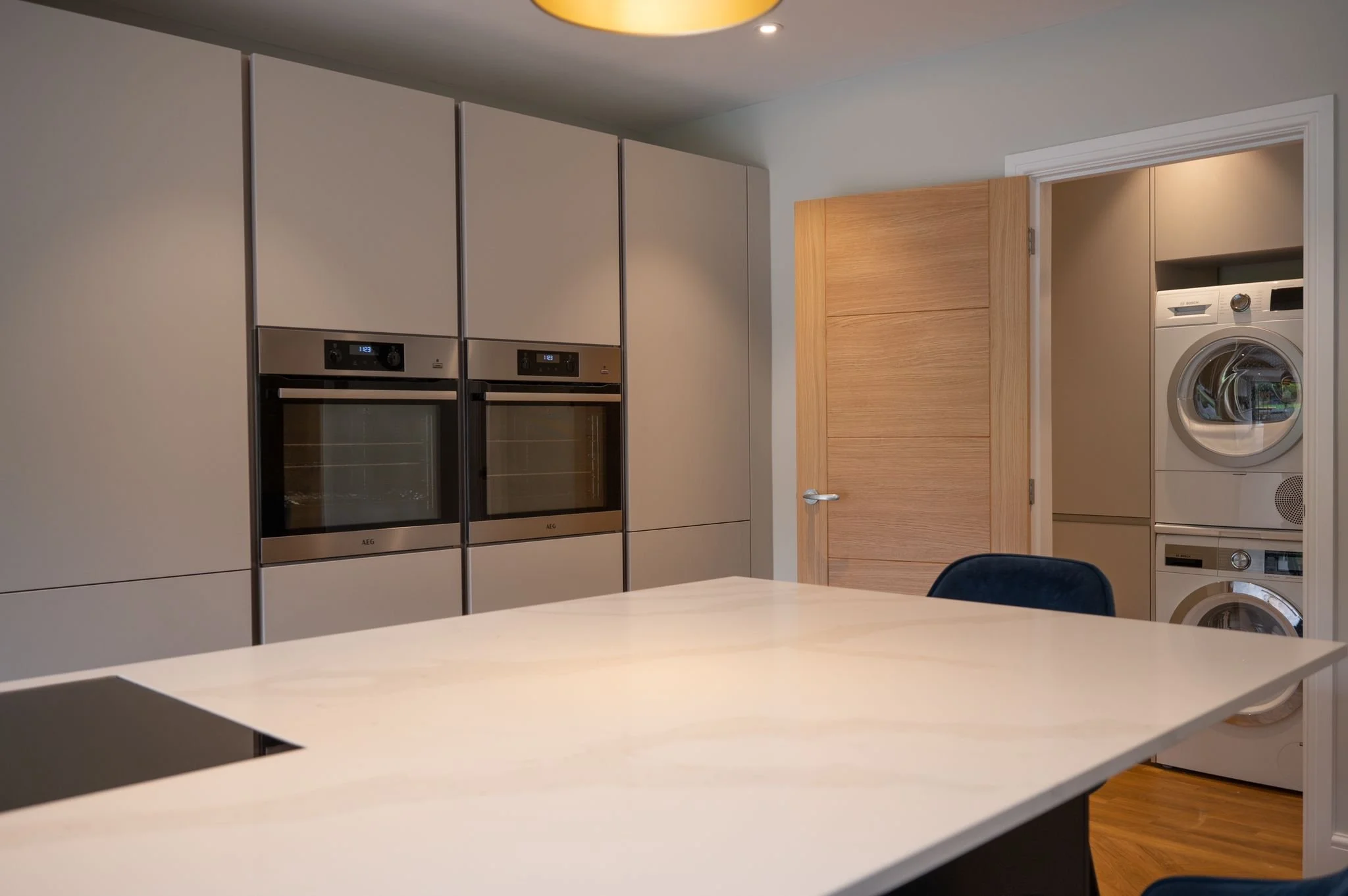 Modern kitchen with built-in double ovens, a white marble countertop, and a laundry area with stacked washer and dryer behind an open wooden door.
