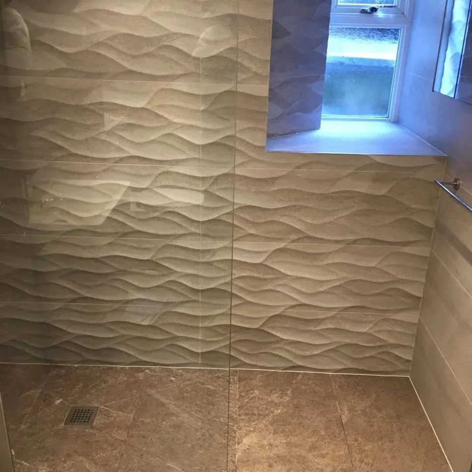 A shower wall with textured beige tiles and a small window, looking down at the tiled floor with a drain.