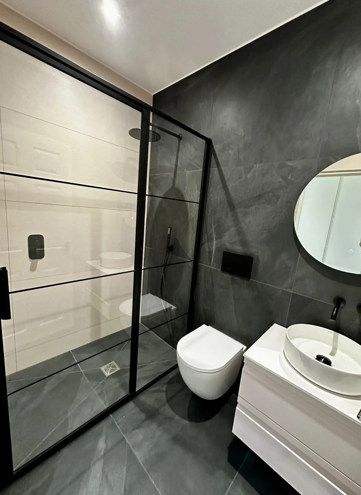 Modern bathroom with dark gray tiled wall and floor, a glass shower enclosure, a white toilet, a white vanity with a vessel sink, and an oval mirror.