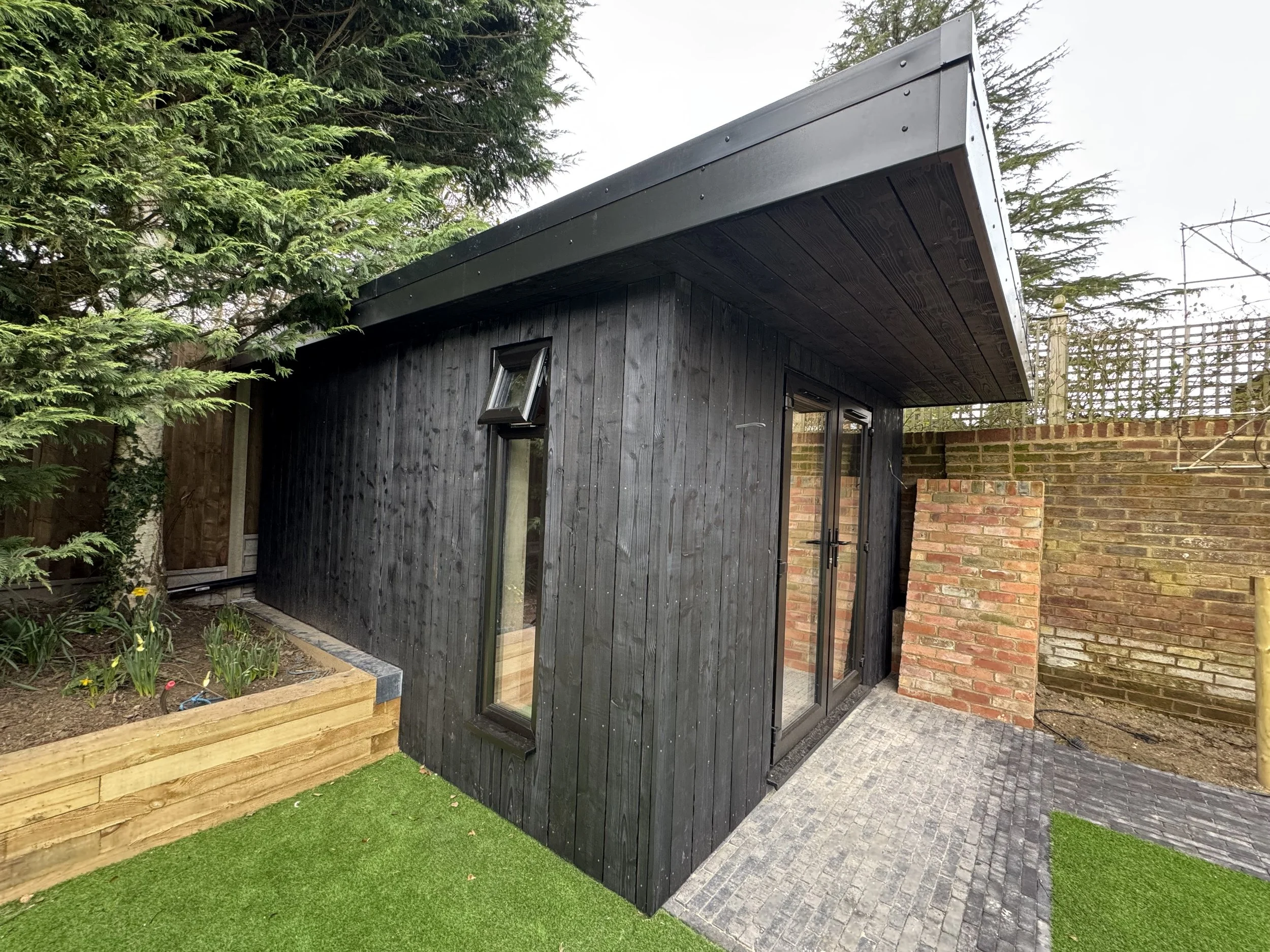 Small modern shed with black wooden exterior, a sliding glass door, and a small window, surrounded by a garden with plants and artificial grass.